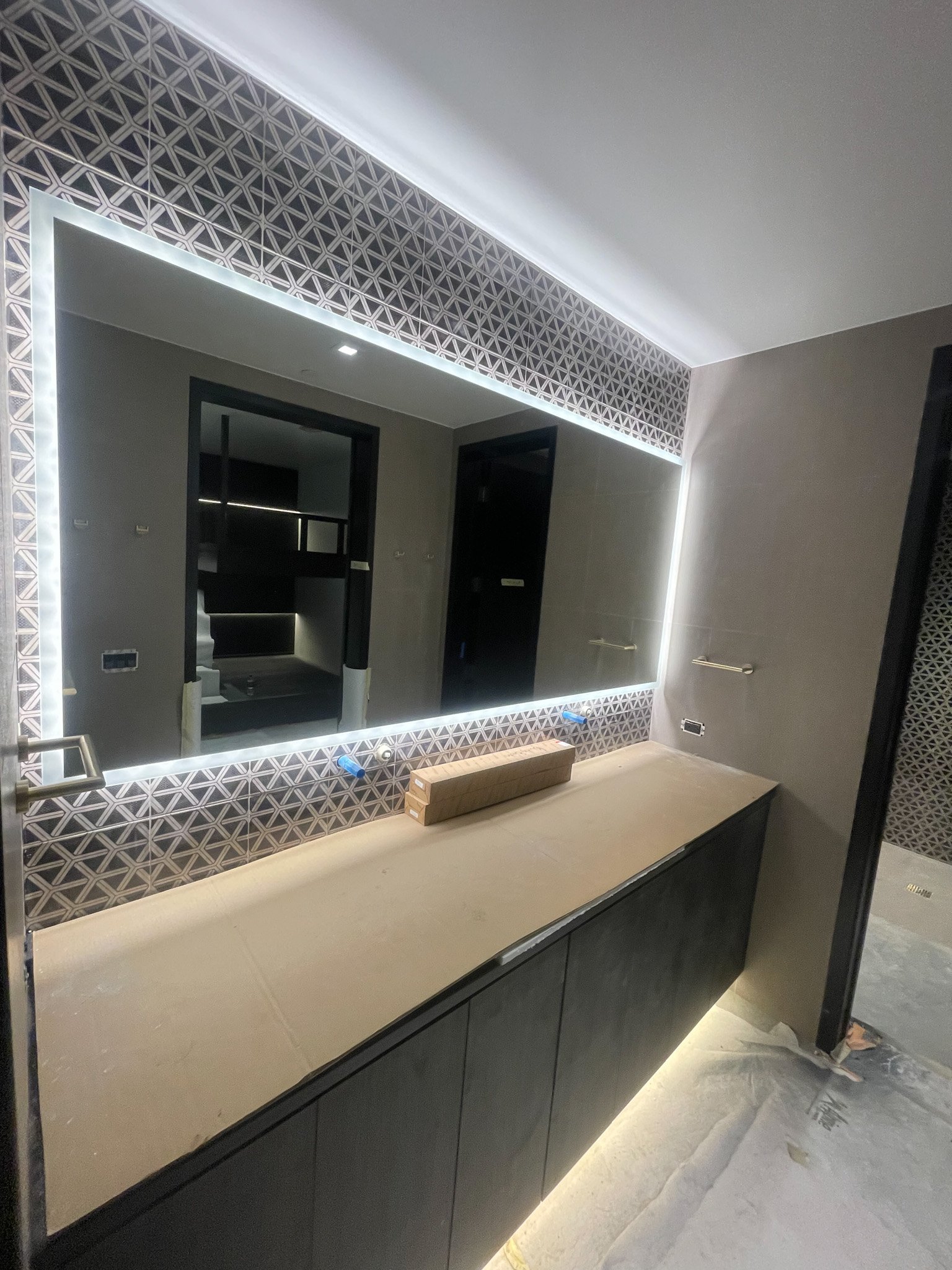 Modern bathroom vanity with a large backlit mirror, black cabinetry, and geometric patterned wall tiles.