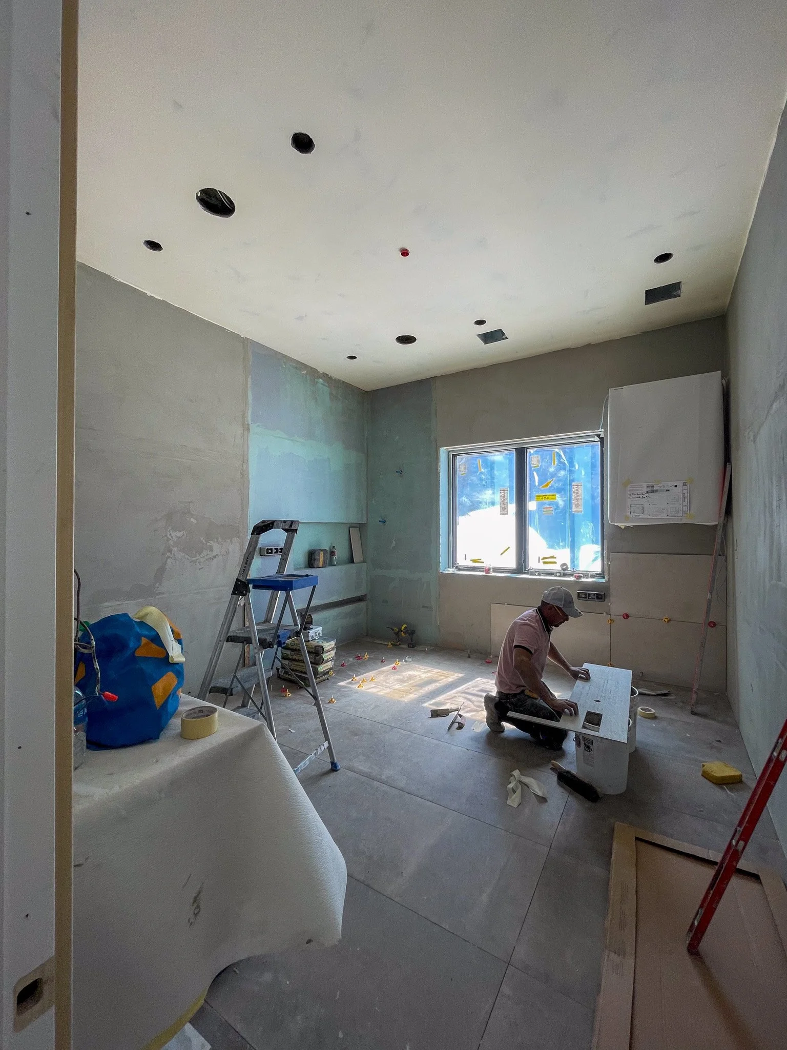 Construction worker kneeling on the floor in an unfinished room with drywall, tools, and ladders, working on the wall or electrical fixtures, with a window letting in natural light.
