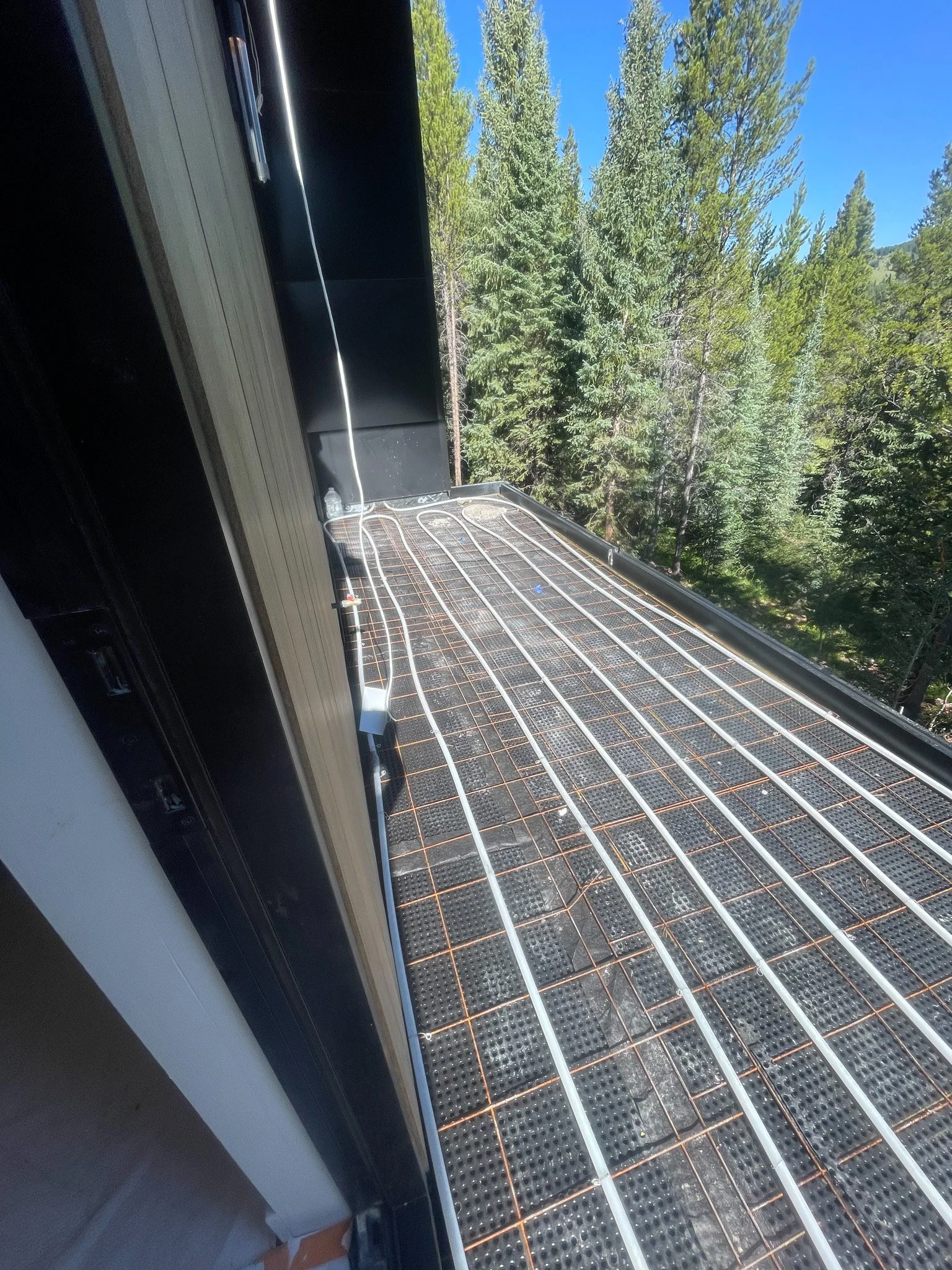 Construction or installation of a heated cable system on a roof with a grid of heating cables and mats, with a view of tall trees and blue sky in the background.