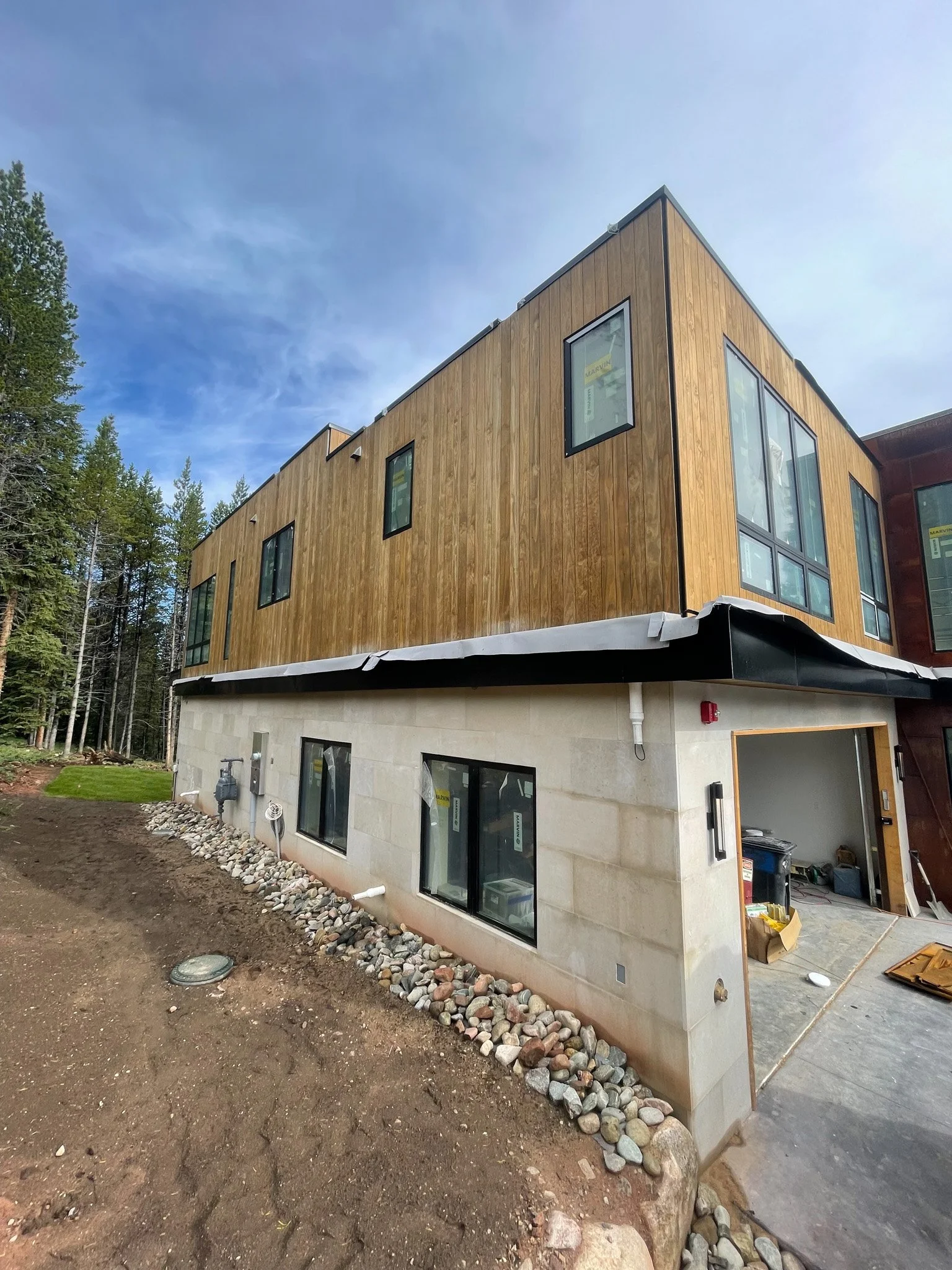 Under construction modern multi-story building with wood and concrete exterior, surrounded by trees and blue sky.