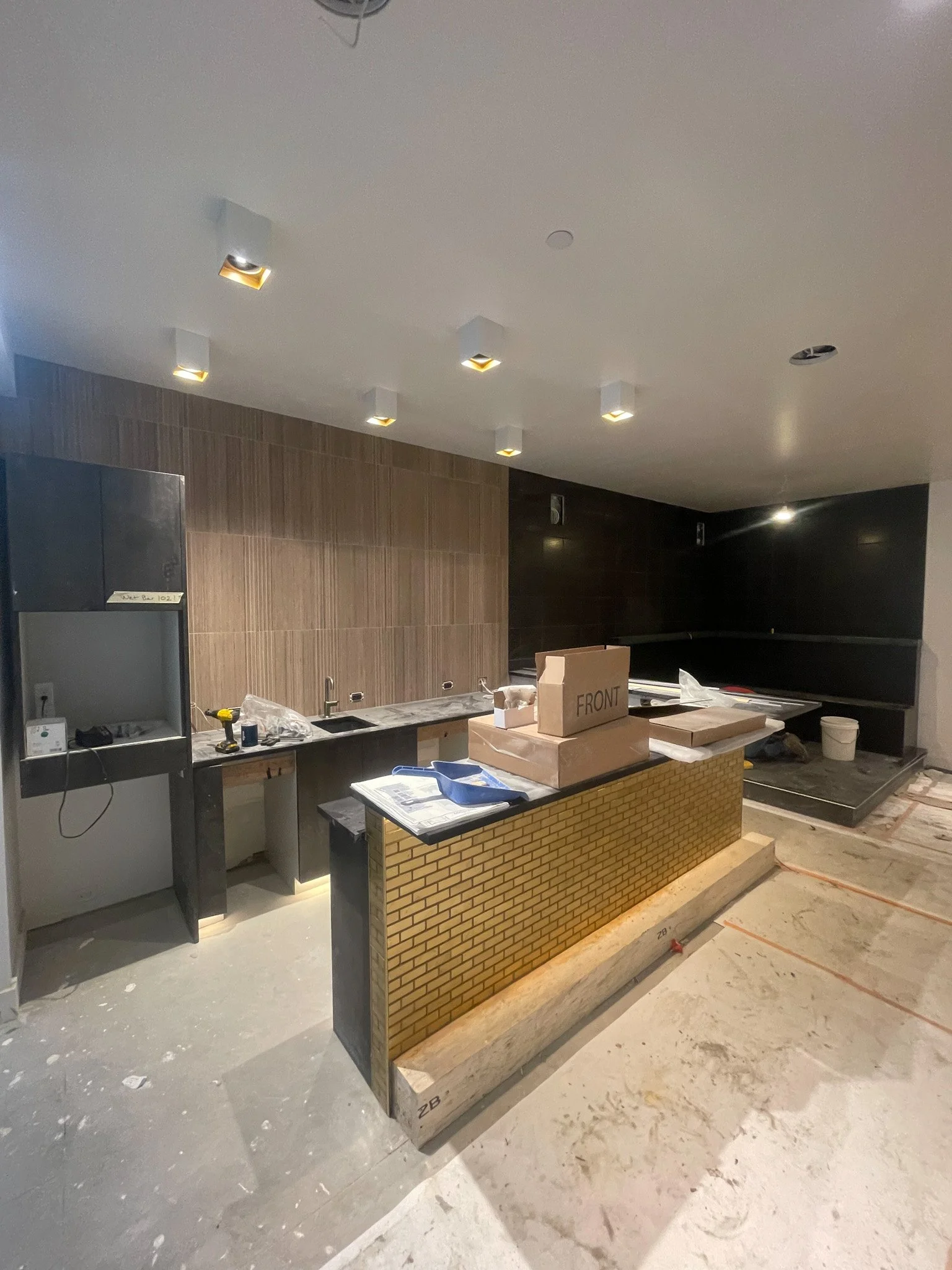 A kitchen under construction with a central island featuring a brick pattern on the front, a black countertop, and unfinished flooring. There are multiple ceiling lights and construction tools and materials scattered around.