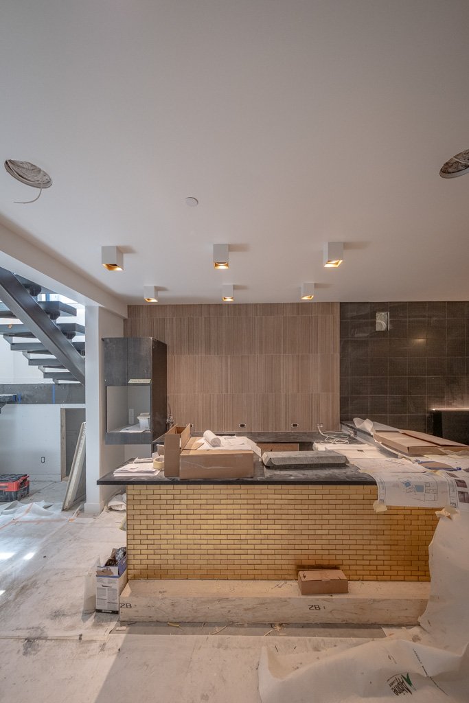 Interior view of a kitchen under construction with a brick island, wooden and dark tile backsplash, and ceiling lights.