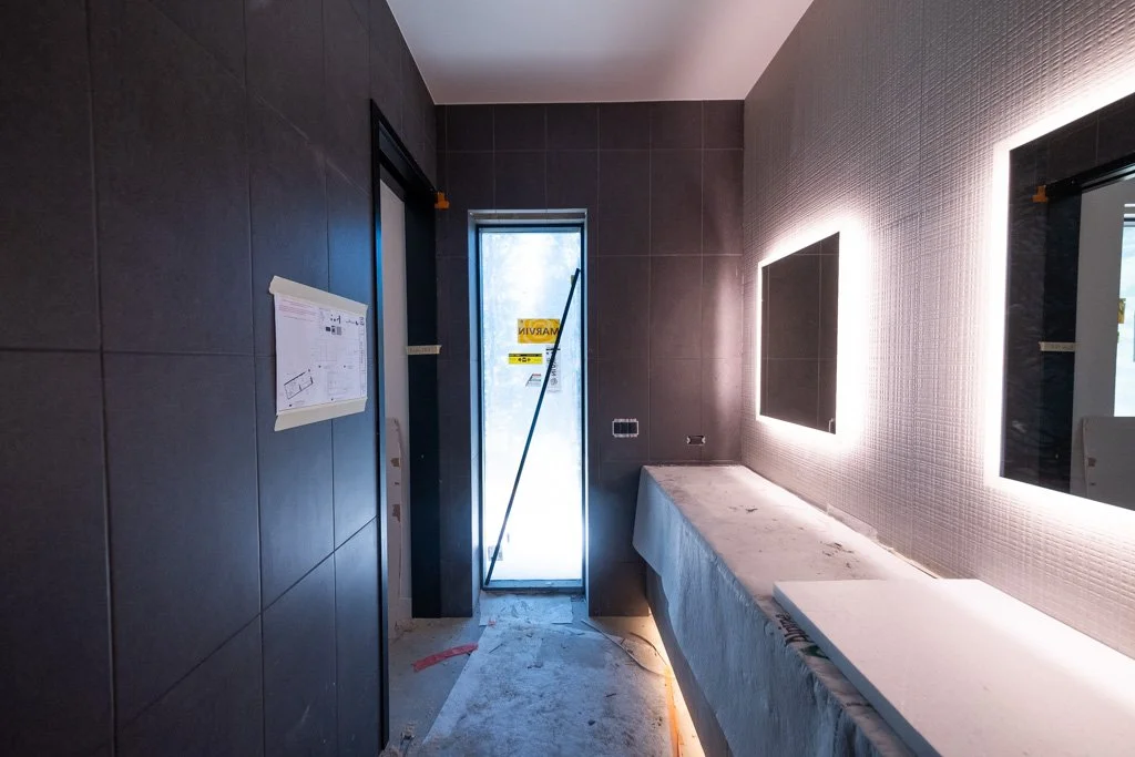 Bathroom under construction with dark tiled walls, a frosted glass door, illuminated mirrors, and a long countertop covered in protective material.