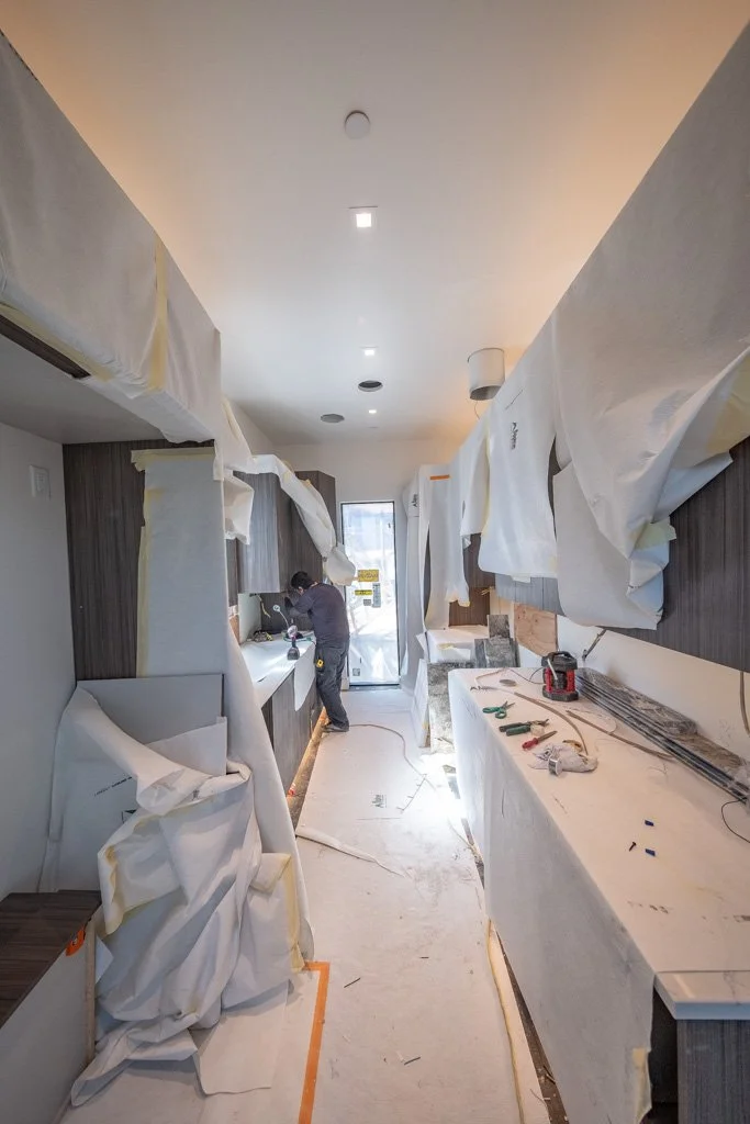 Construction workers remodeling a kitchen with what appears to be white protective sheets hanging from the ceiling and walls, tools on the counter, and a person working near the sink with a door at the back leading outside.