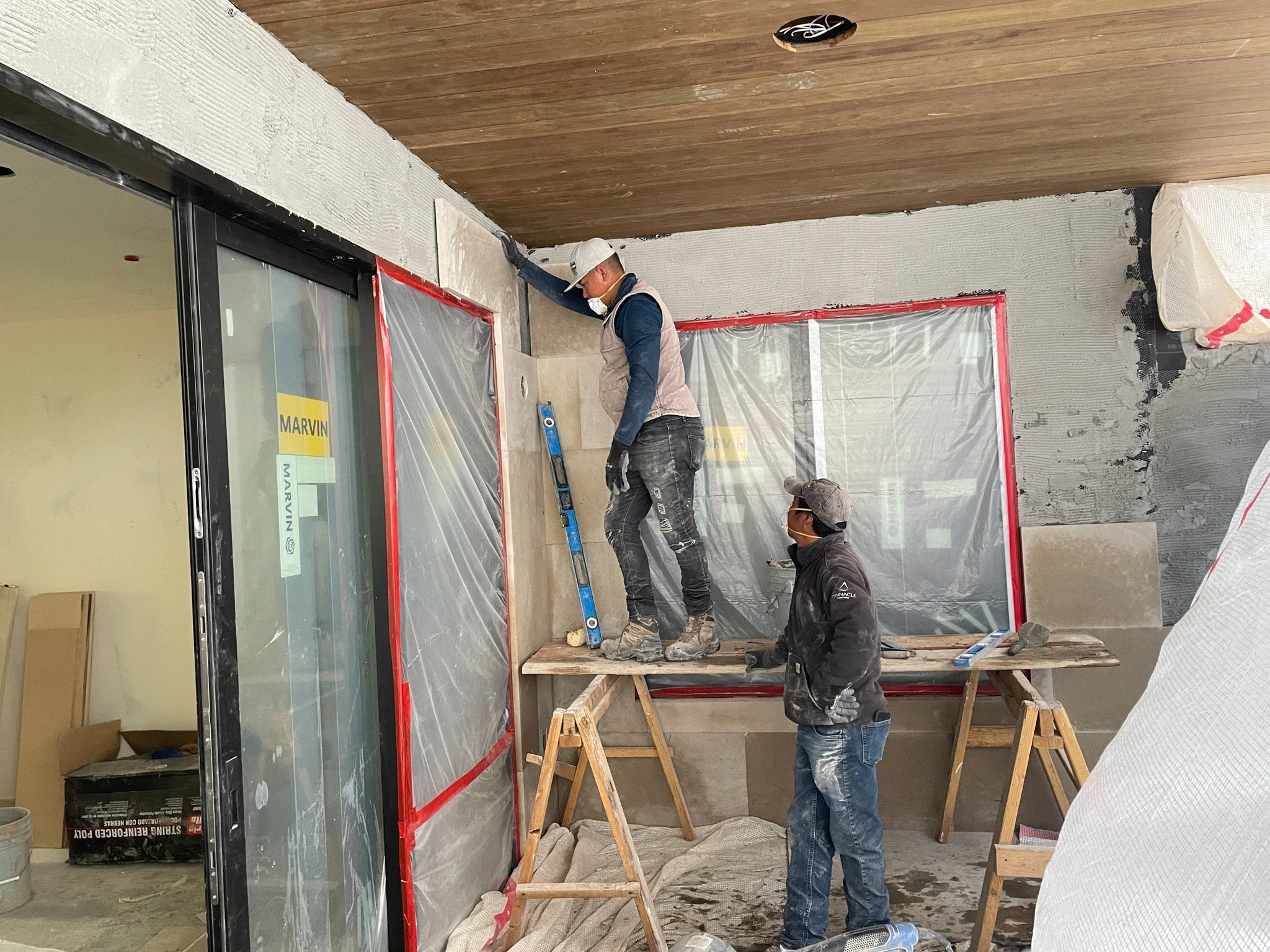Two construction workers installing ceramic tiles on a wall, with plastic sheeting covering the window area for protection.