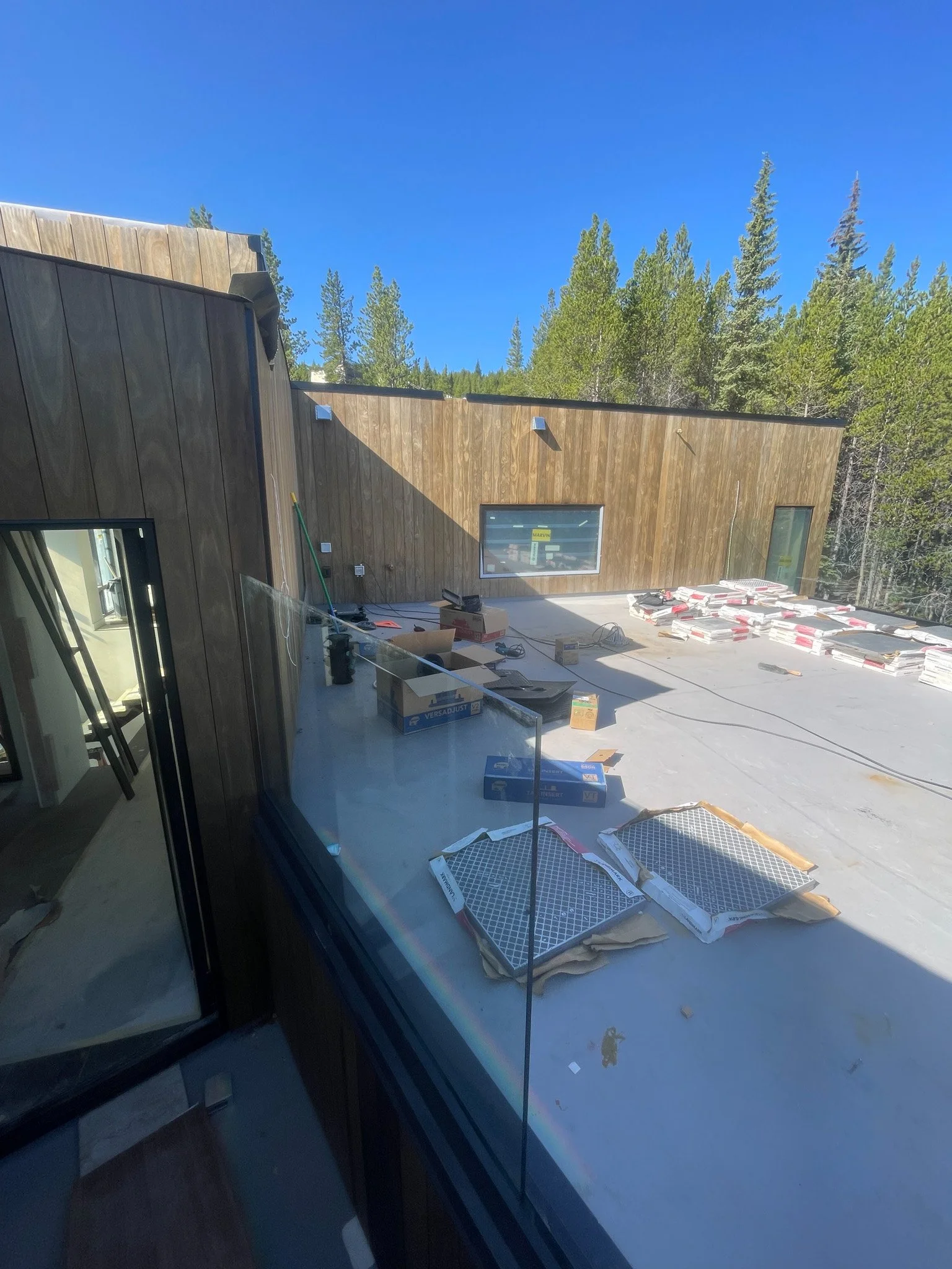 Construction site on the roof of a building with various construction materials and tools, surrounded by trees under a clear blue sky.