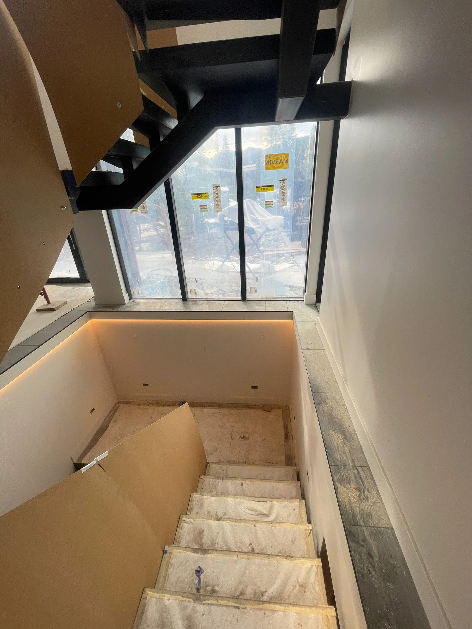 Interior view of a staircase in a building under construction, with protective coverings on the steps, a large glass window at the top, and some construction signs on the window.
