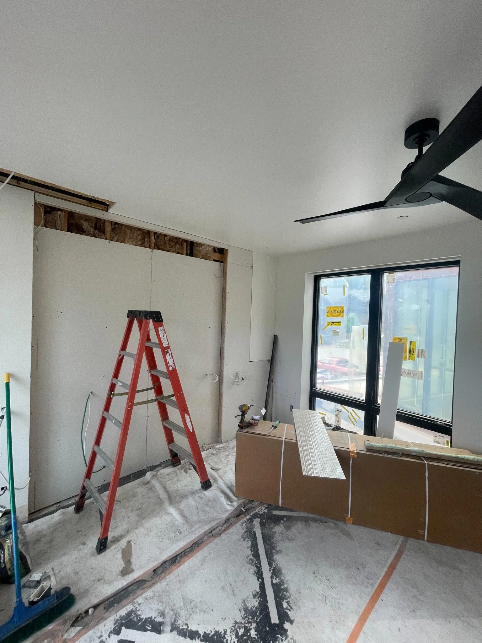 Room under renovation with a red step ladder, construction tools, and materials, large window, ceiling fan, and unfinished wall.