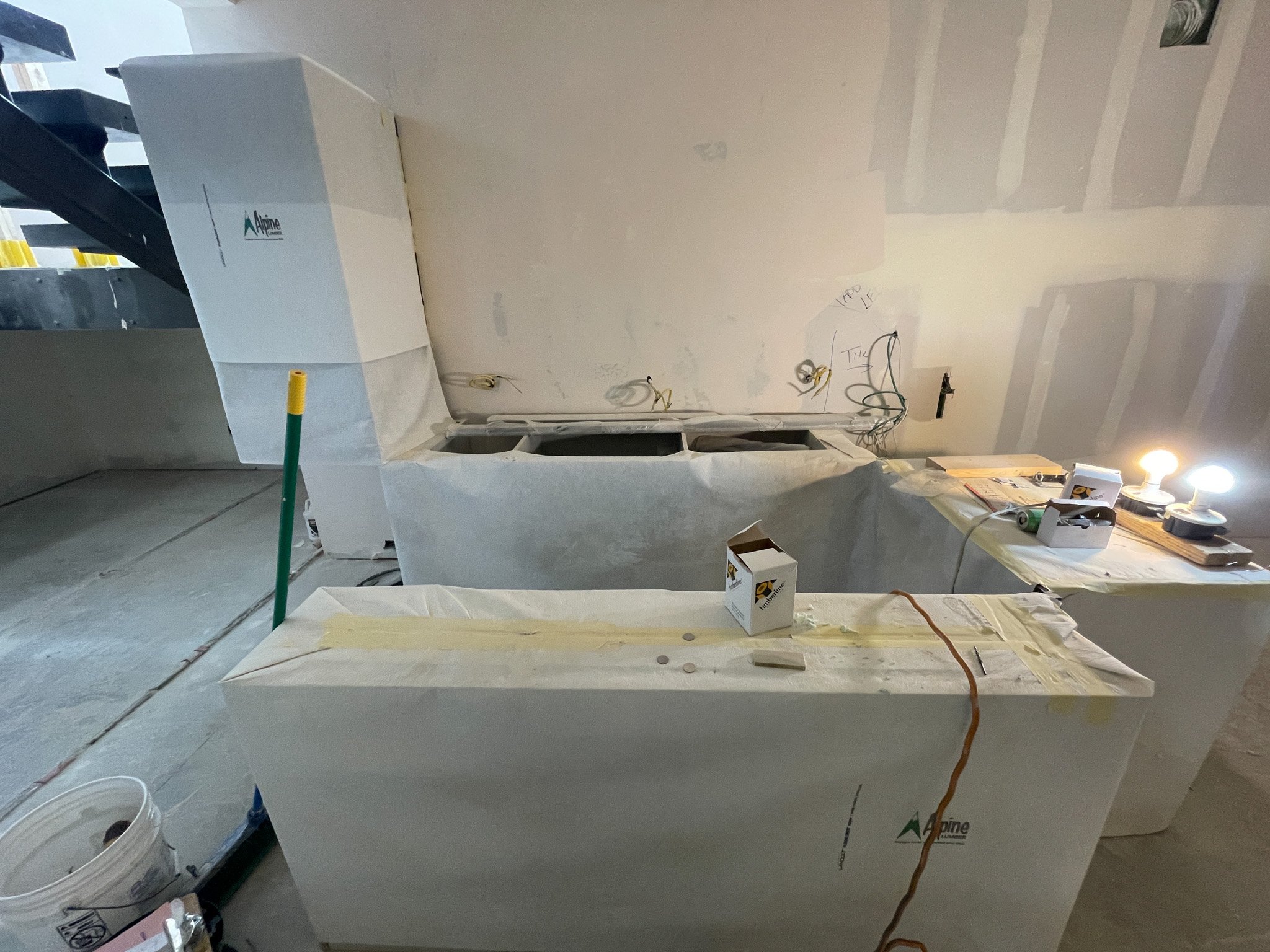 Construction site with drywall installation, covered ventilation system, and work lights, showing unfinished walls and construction materials.