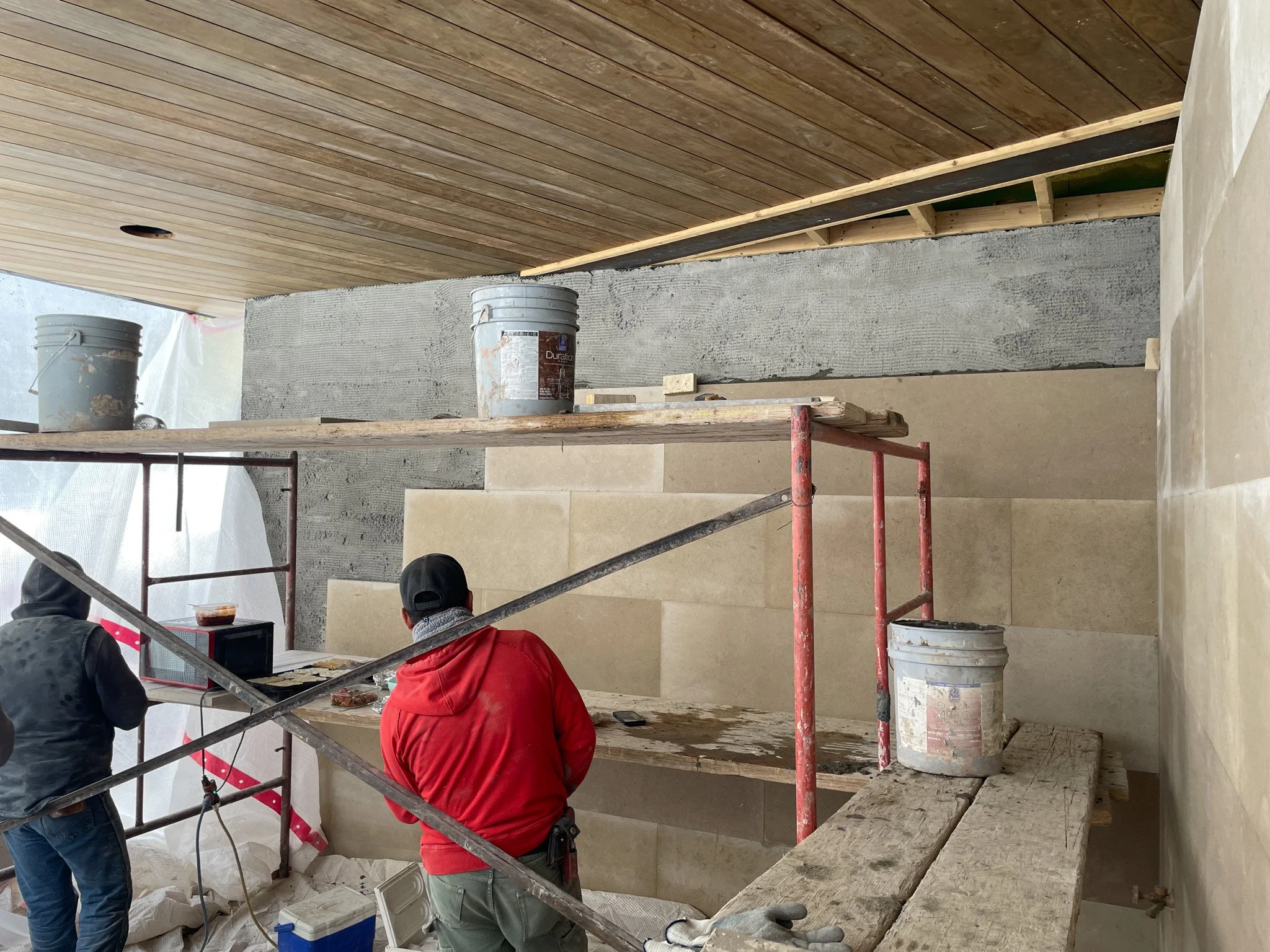 Construction workers working on wall installation with scaffolding, buckets, and construction tools in an indoor space.