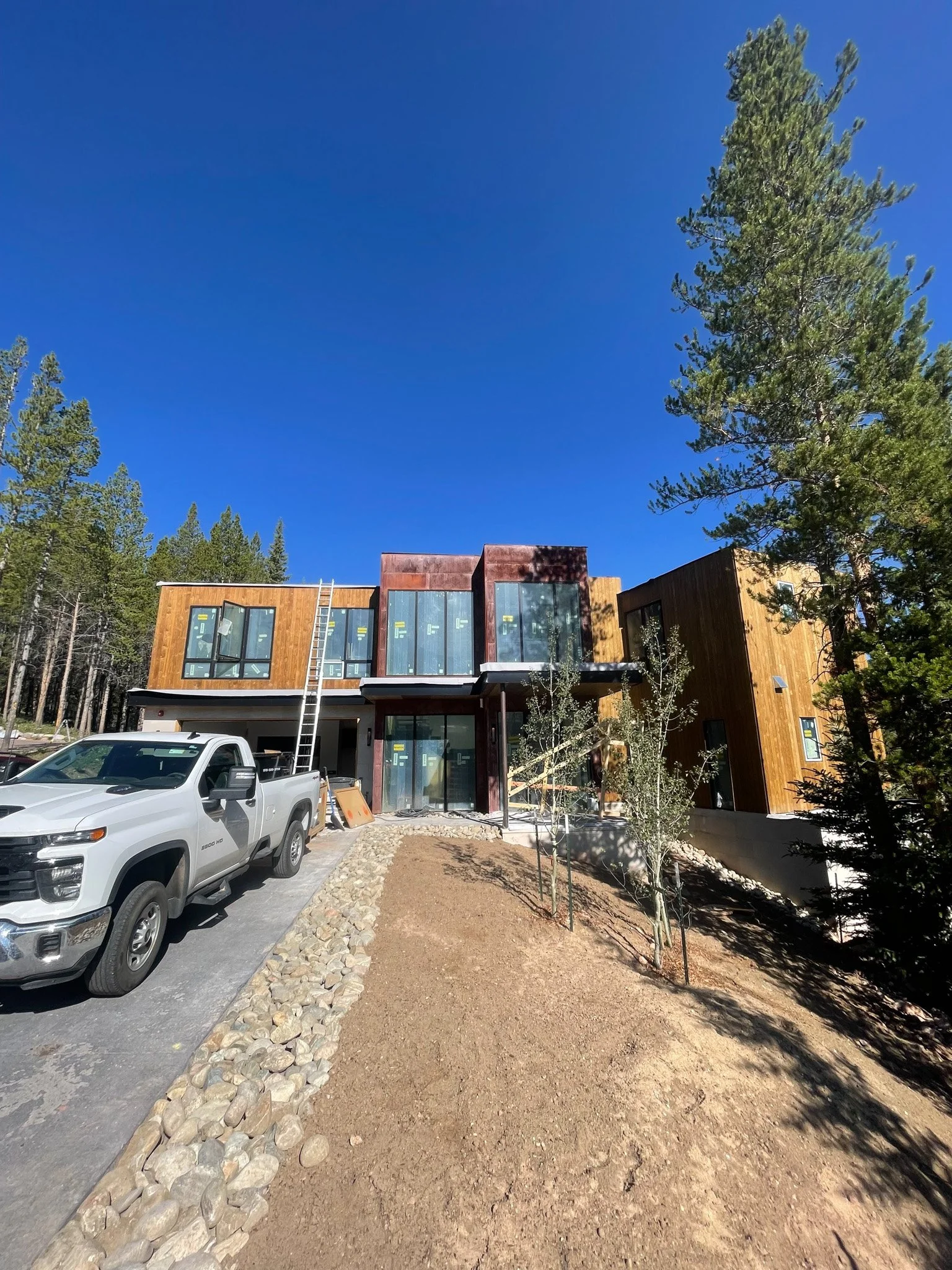 Under construction modern house with wooden and glass exterior, parked white pickup truck, construction ladder, and bare soil in a wooded area with tall trees under a clear blue sky.