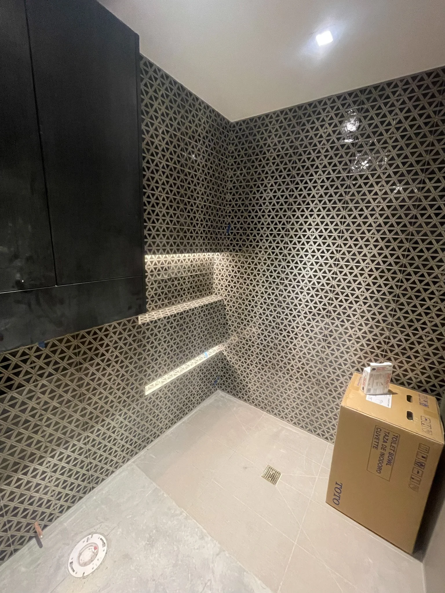 A partially finished bathroom with patterned wall tiles, a black cabinet, and a cardboard box on the floor.