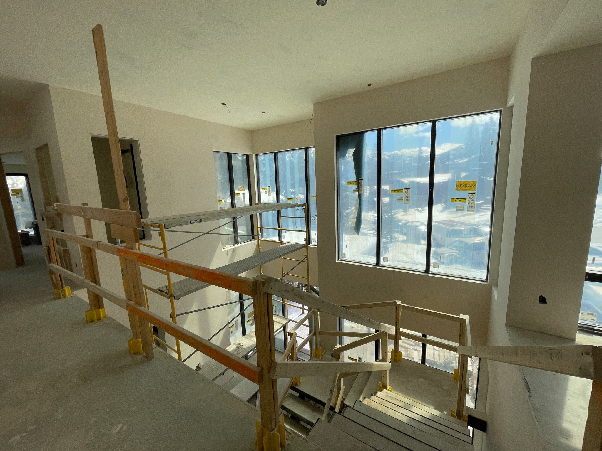 Interior of a building under construction featuring a staircase with temporary wooden railings, large windows with stickers, and sunlight coming through the windows.