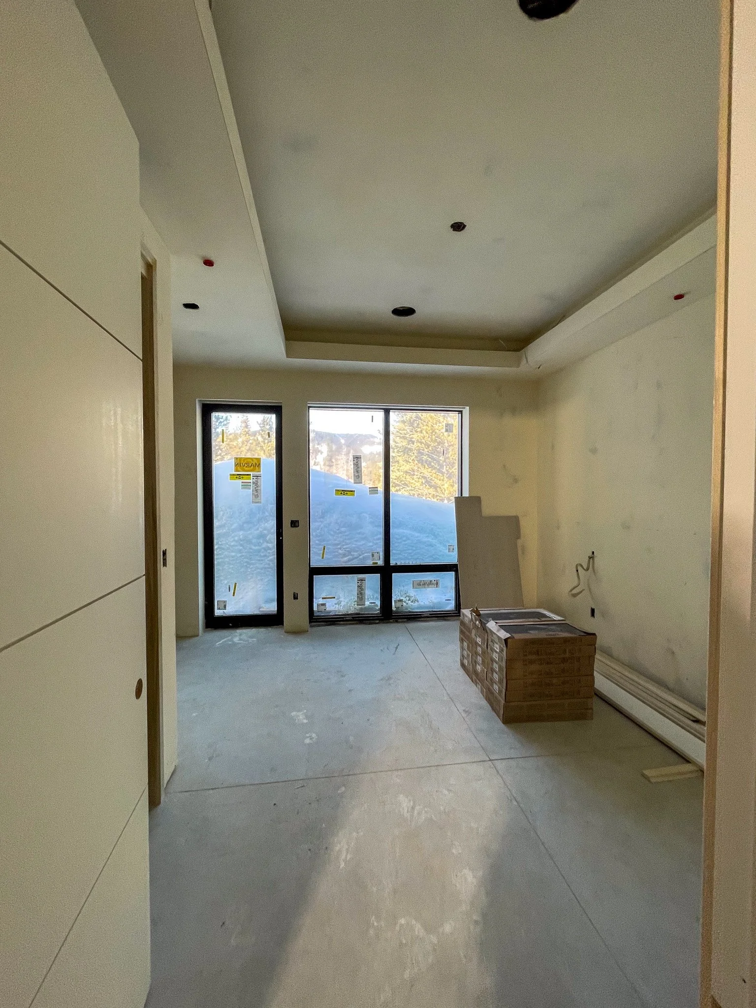 Interior of a room under construction with large window/door, construction materials on the floor, and unfinished walls.