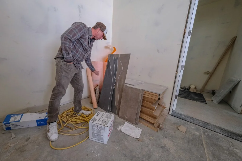 Man at a construction site looking at building materials and tools, with loose wiring on the floor and various tiles and wood pieces stacked against the wall.