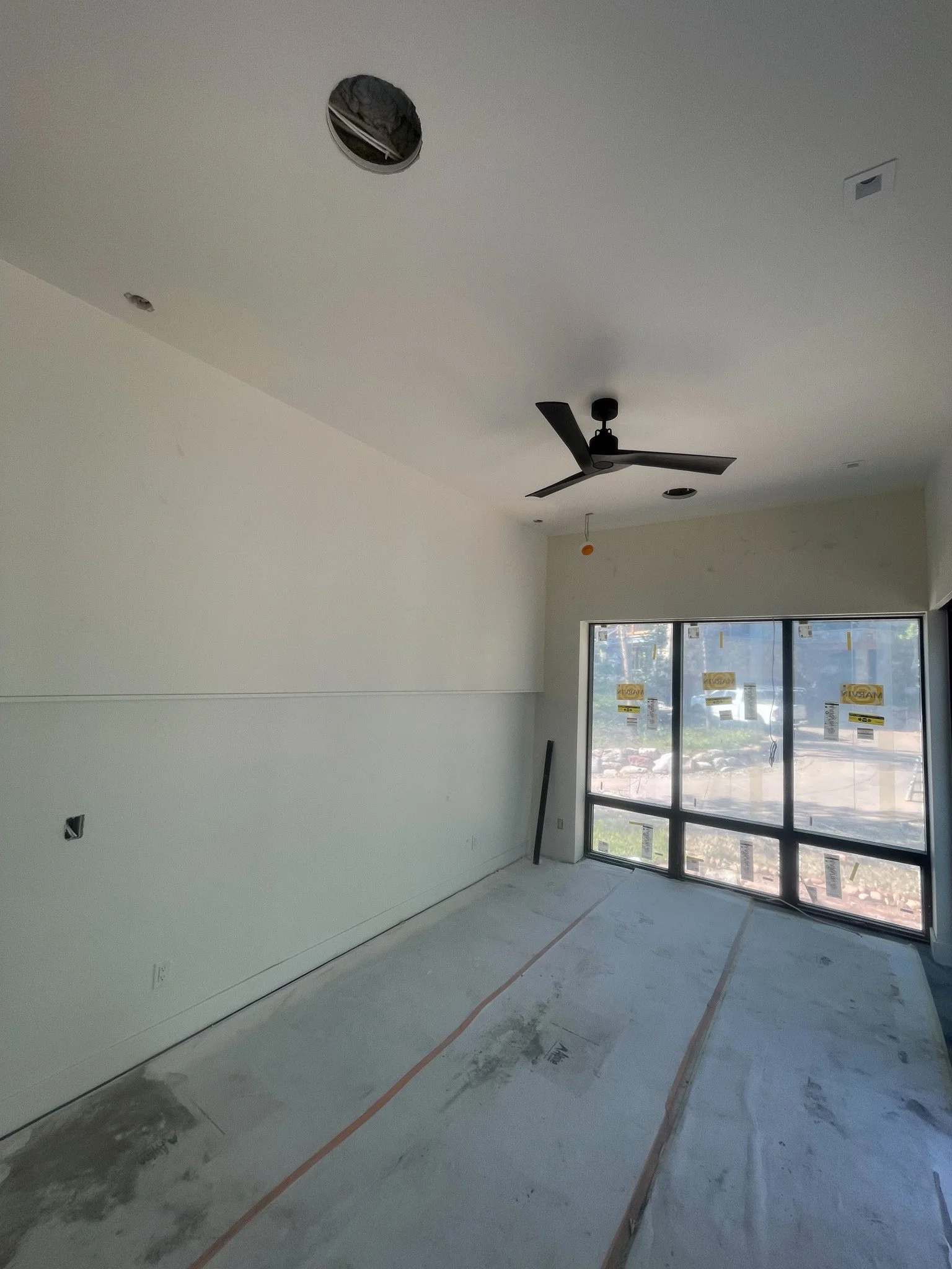 Interior of a room under construction with a white wall, large window, ceiling fan, and unfinished ceiling with a hole for lighting installation.