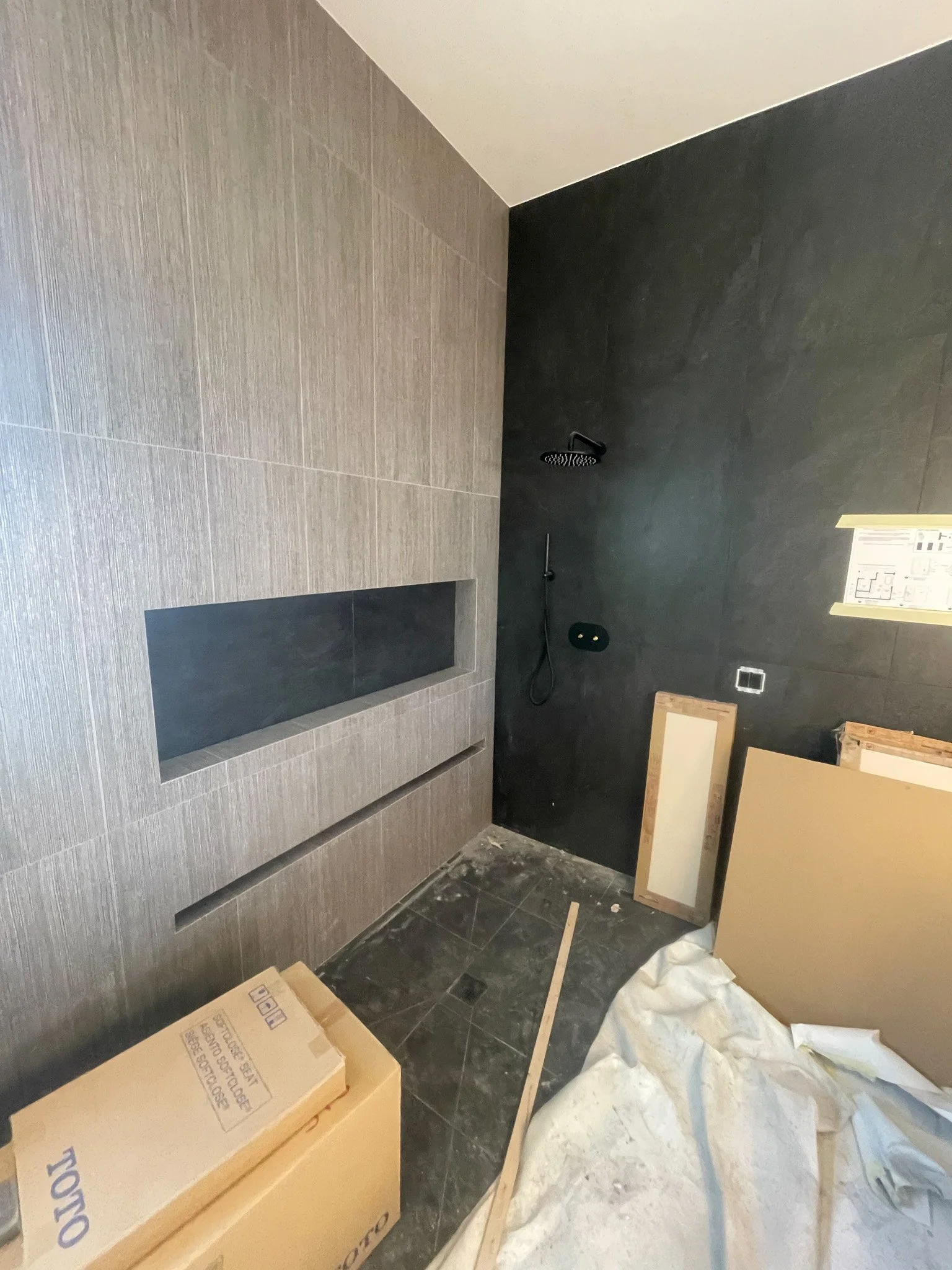 Interior of a bathroom under construction with dark tiled shower area, black shower head, and a niche in the wall. Construction materials and debris are on the floor.