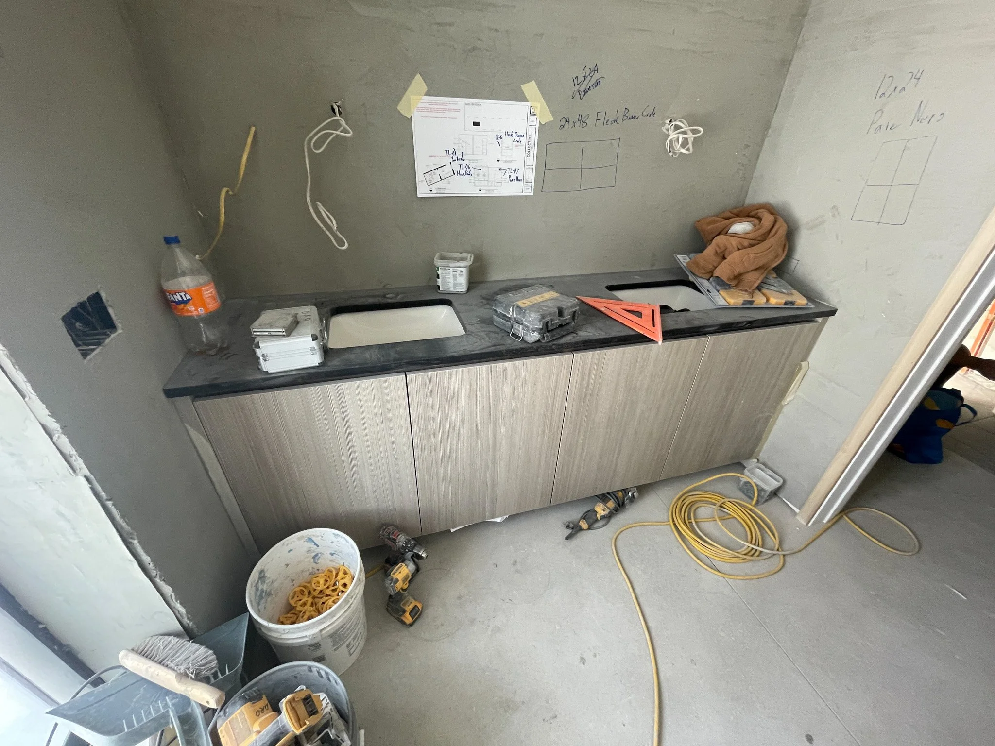A construction site kitchen with a countertop, cardboard cabinets, and construction tools including drills, measuring tools, and extension cords. There are handwritten notes and diagrams on the walls.