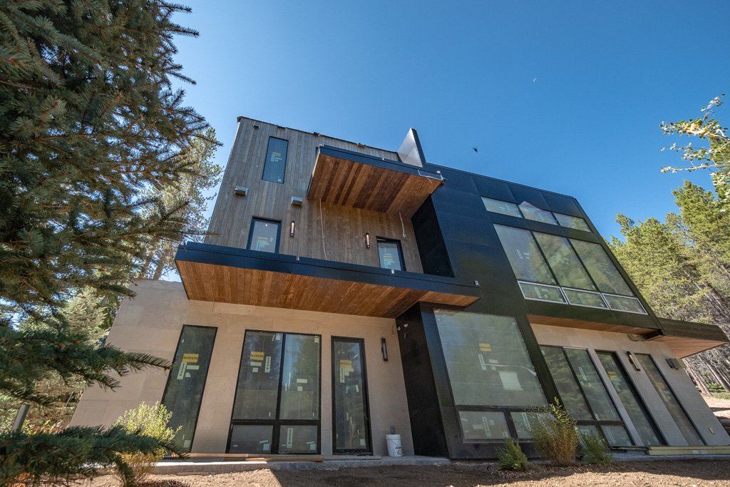Modern multi-story house with large glass windows, wooden and concrete exterior, situated among trees under a blue sky.