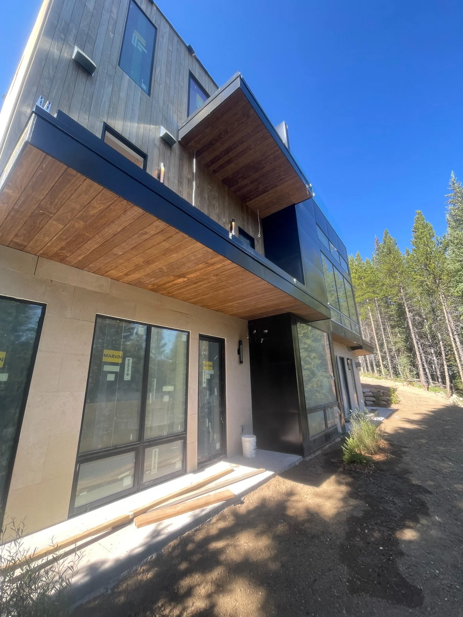 A modern multi-story house under construction with large glass windows, wooden panels, and a balcony, surrounded by trees and a clear blue sky.