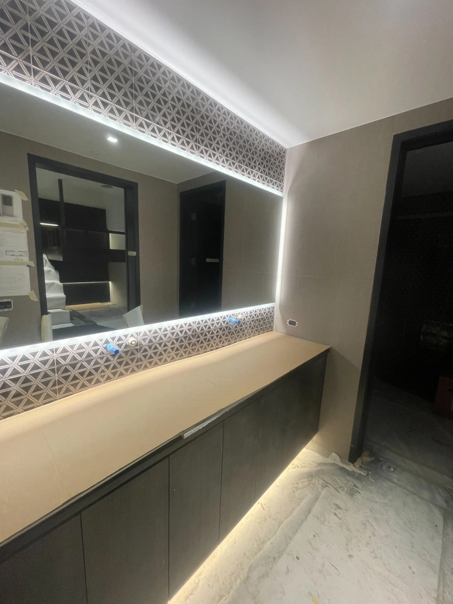 Modern bathroom vanity with a large mirror, backlit with LED lights, featuring geometric patterned tiles above the mirror and under-cabinet LED lighting. The vanity has a beige countertop and dark storage cabinets below, with construction tools and d