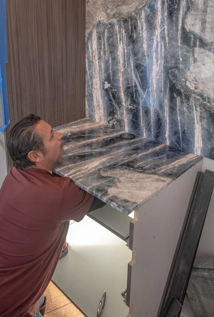 A man with slicked-back hair leaning on a marble countertop in a modern kitchen or bathroom, with a marble backsplash behind him.