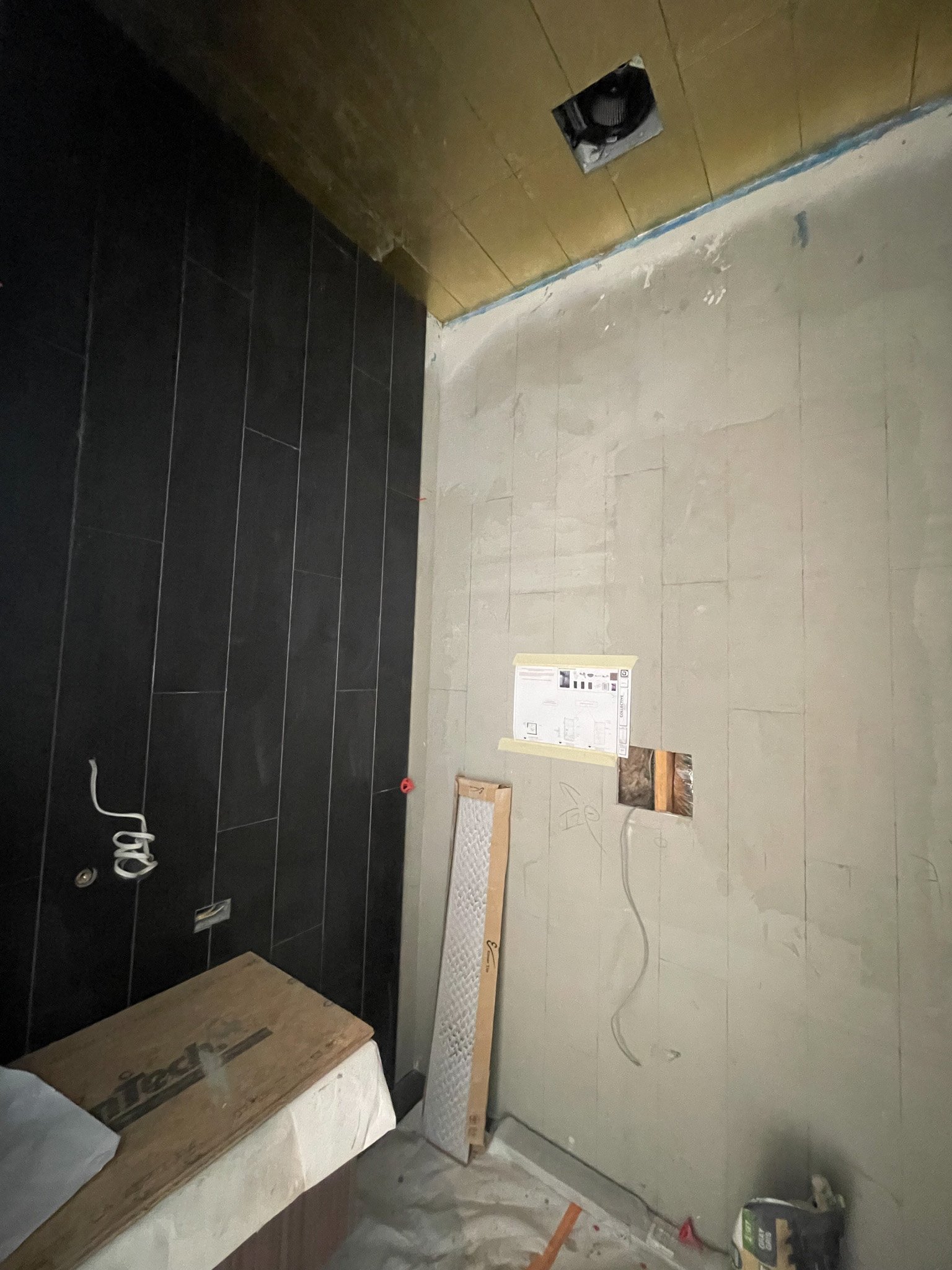 Partially completed bathroom with black tile wall, unfinished gray wall with electrical wiring, a vent opening, and construction materials on the floor.