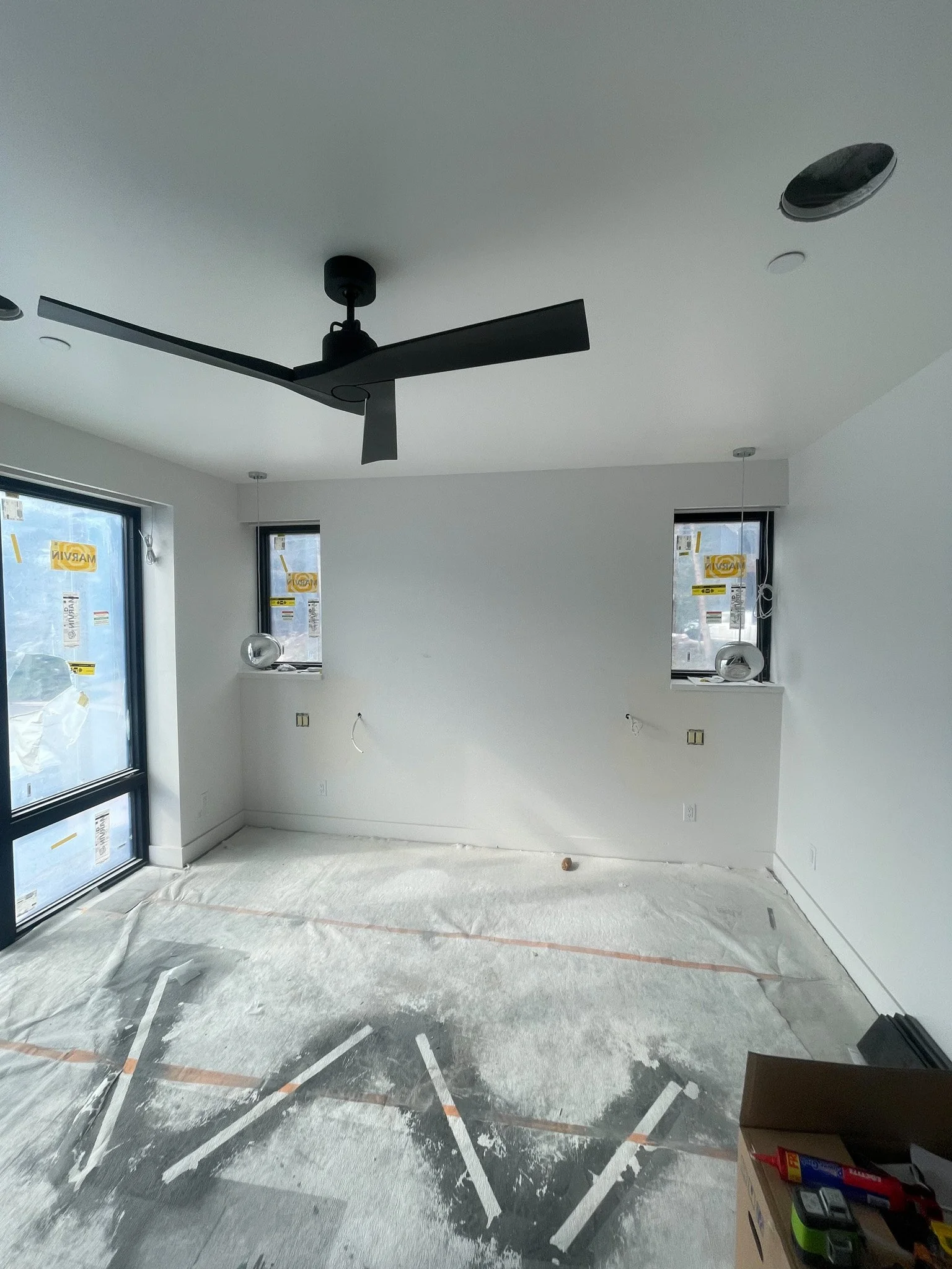 Empty room under construction with white walls, two windows, a black ceiling fan, and some construction materials on the floor.