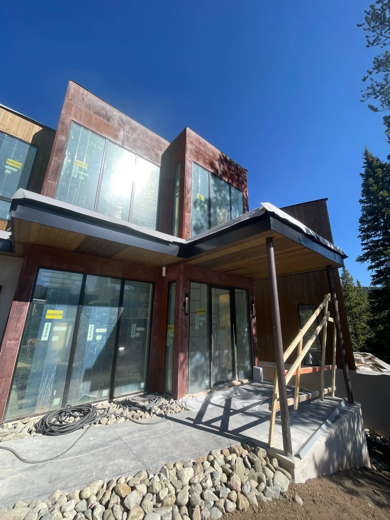 Under-construction modern house with large glass windows, wooden and metal exterior, and a small covered porch with a ramp, surrounded by trees and clear blue sky.
