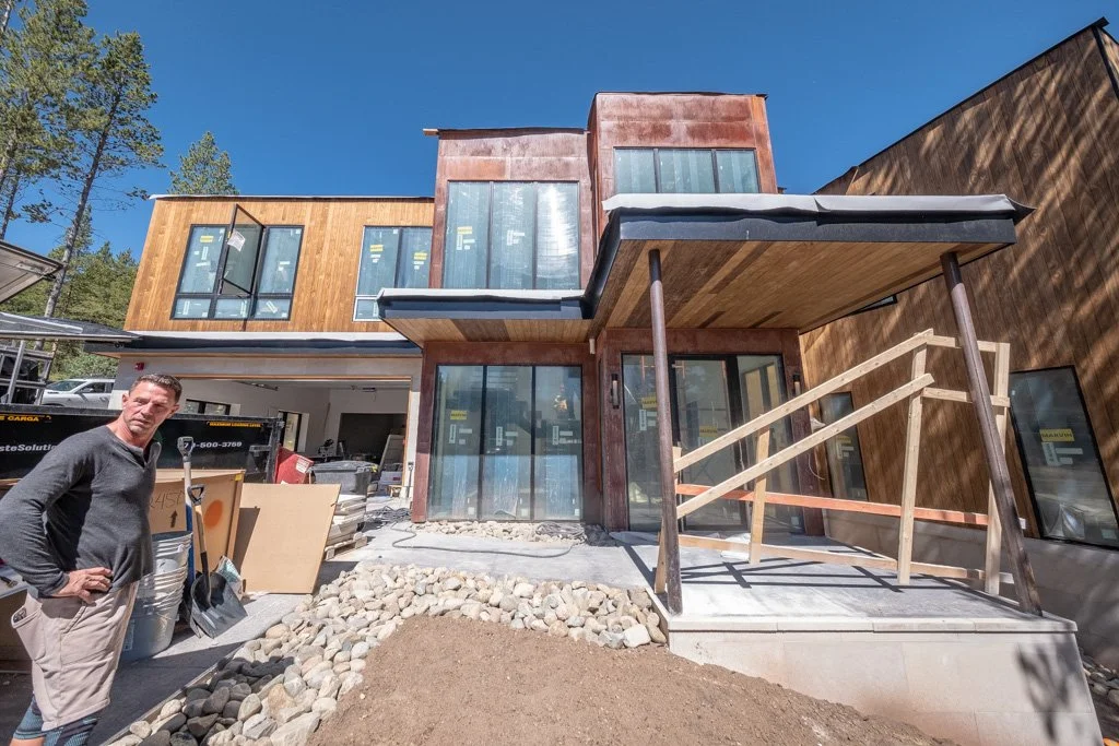 Man standing outside under construction of a modern house with wooden and rust-colored metal siding, large windows, and a small elevated porch with wooden railing and support posts.