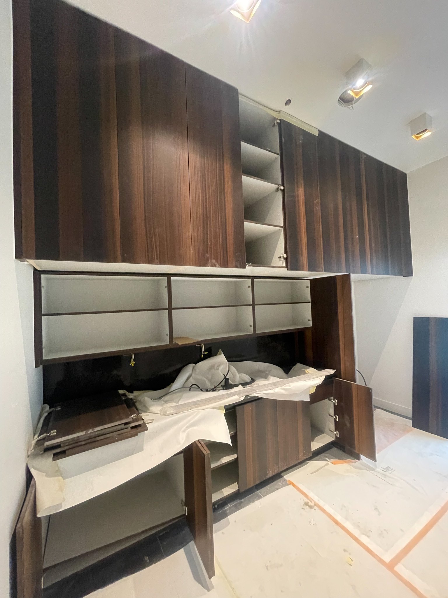 Kitchen cabinet installation in progress with open doors and shelves, some materials covered with paper, in a room with tile floor and installed ceiling lights.