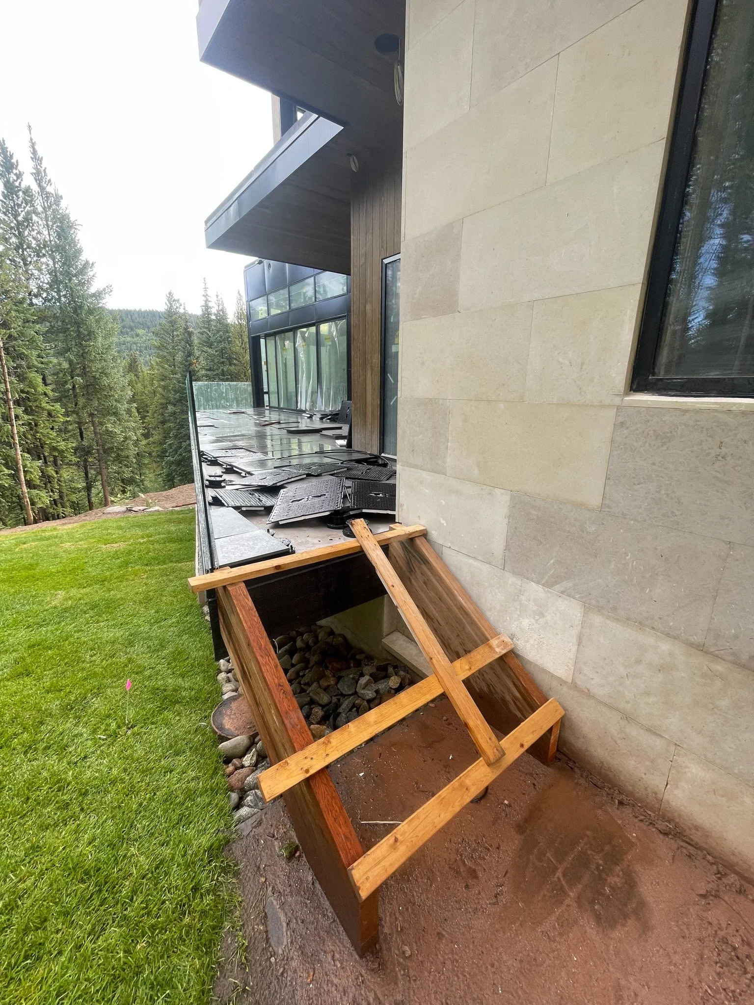 Unfinished wooden planter box with rocks inside, situated on dirt area near the exterior wall of a modern house under construction, with a hillside and forest in the background.