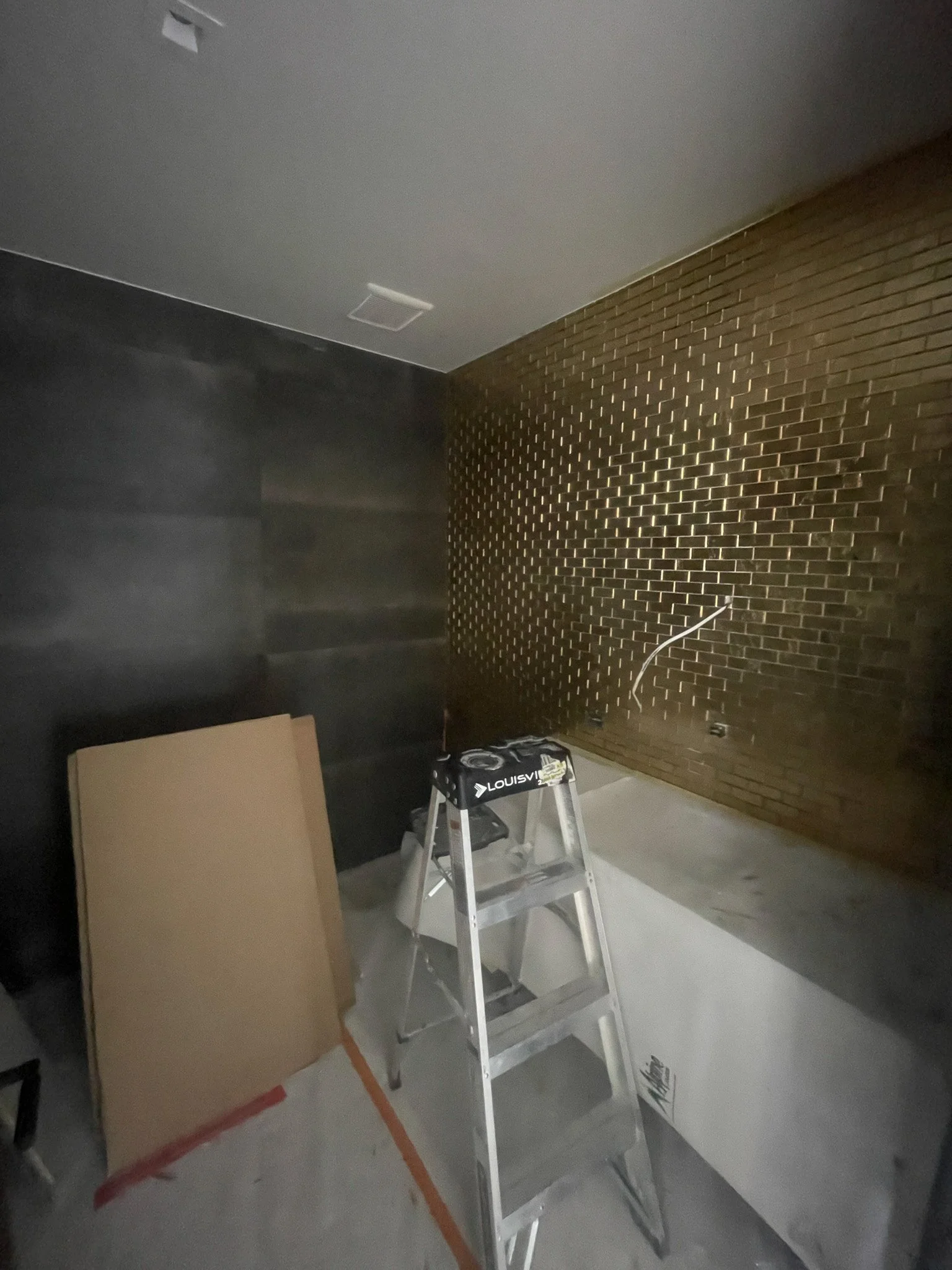 Interior of a room under renovation with a stepladder, cardboard panels, and a black wall adjacent to a brick wall.