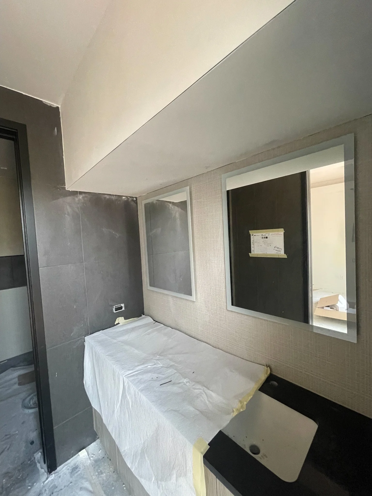 Bathroom under construction with a black countertop, a white undermount sink, two mirrors on the wall, and protective paper covering the sink.
