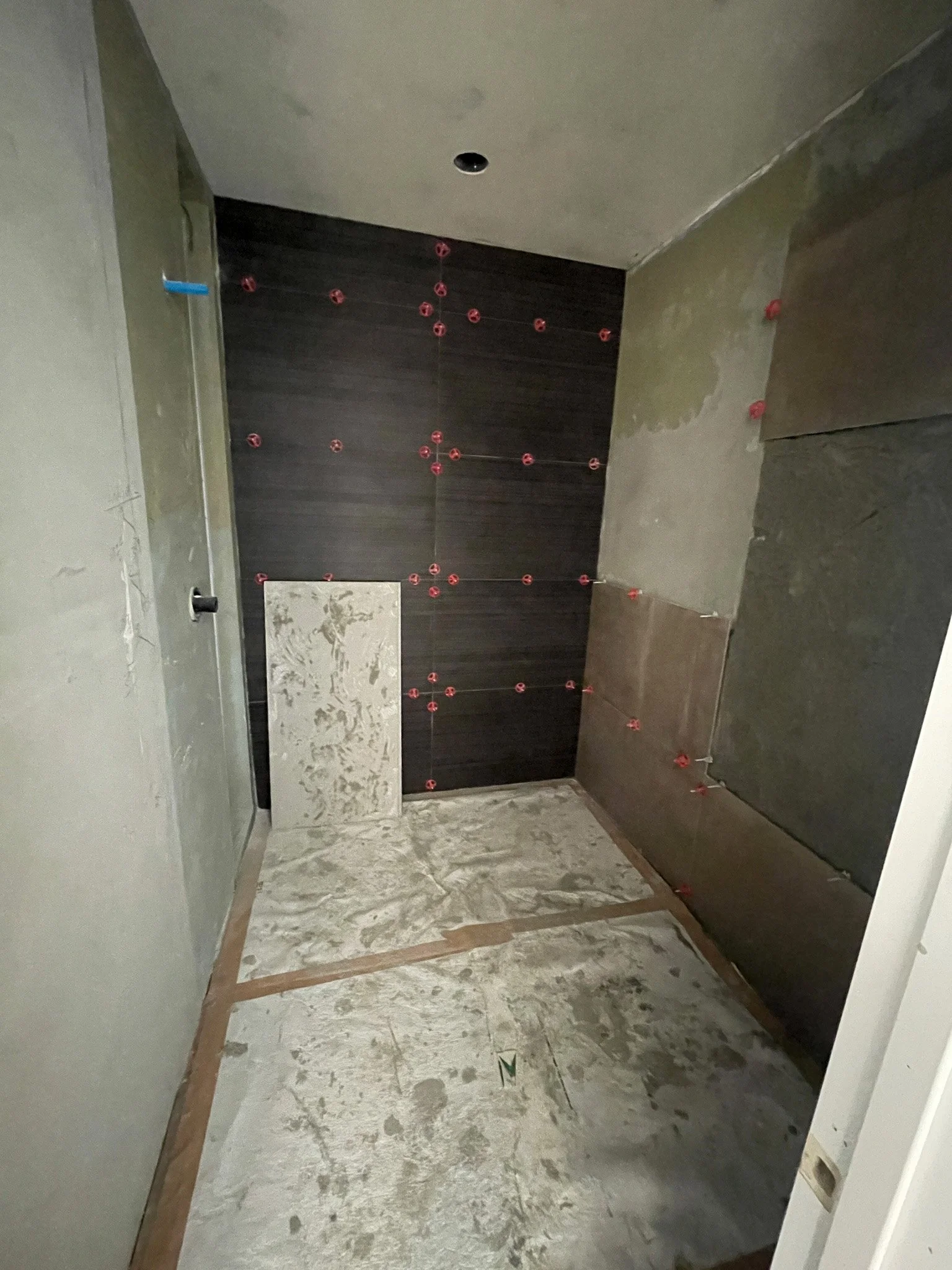 Bathroom under construction with dark brown wall tiles on the back wall, and a partially installed floor tile leaning against the wall. Red spacers are visible between tiles, and the floor is unfinished.