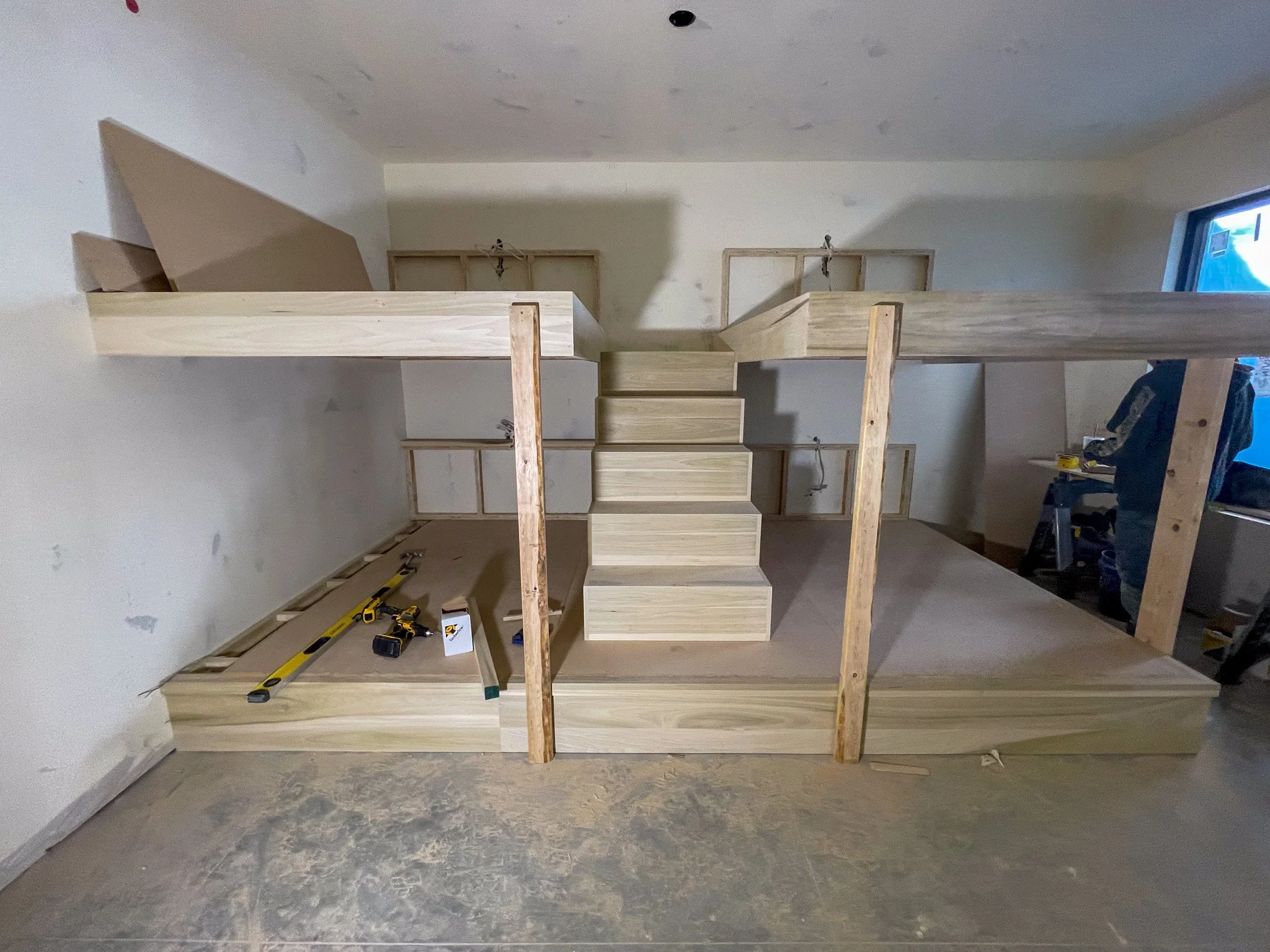 Unfinished wooden staircase with two levels under construction, surrounded by construction tools and materials in a room with unpainted walls.