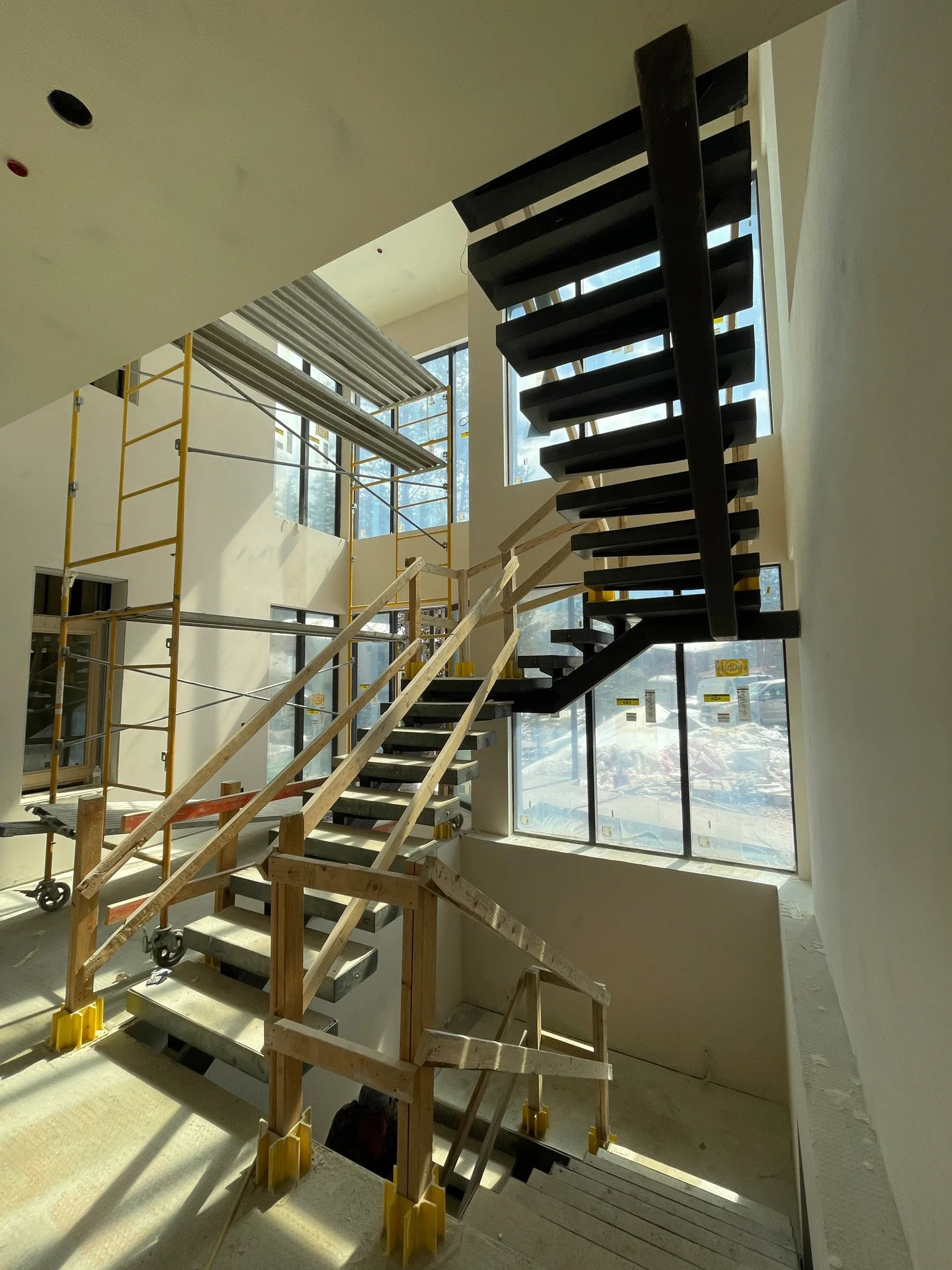 Interior view of a building under construction, showing stairs, scaffolding, and large windows.