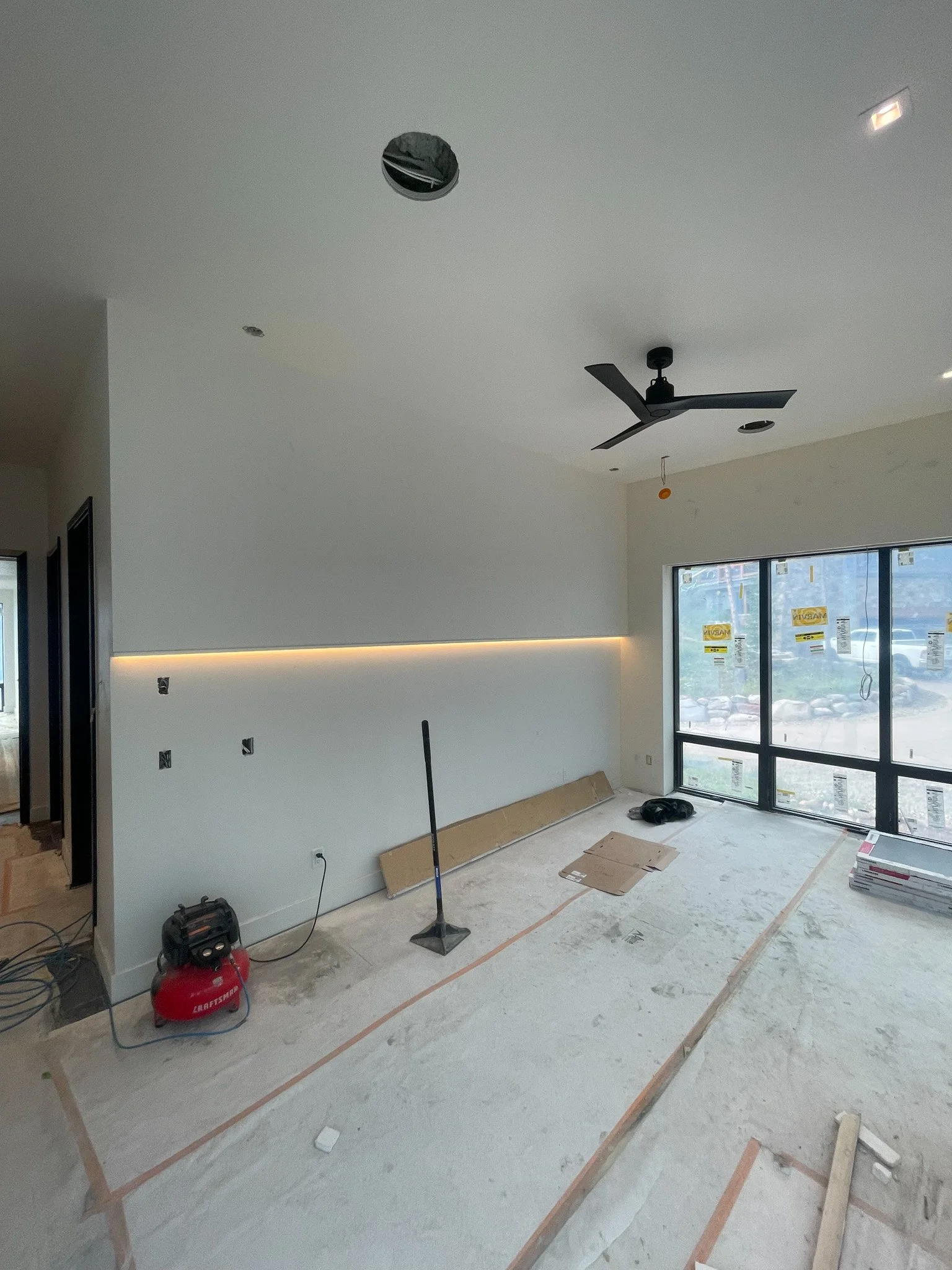 Interior of a room under construction with a large window, a black ceiling fan, and a partially installed ceiling light fixture, with construction tools and materials on the floor.