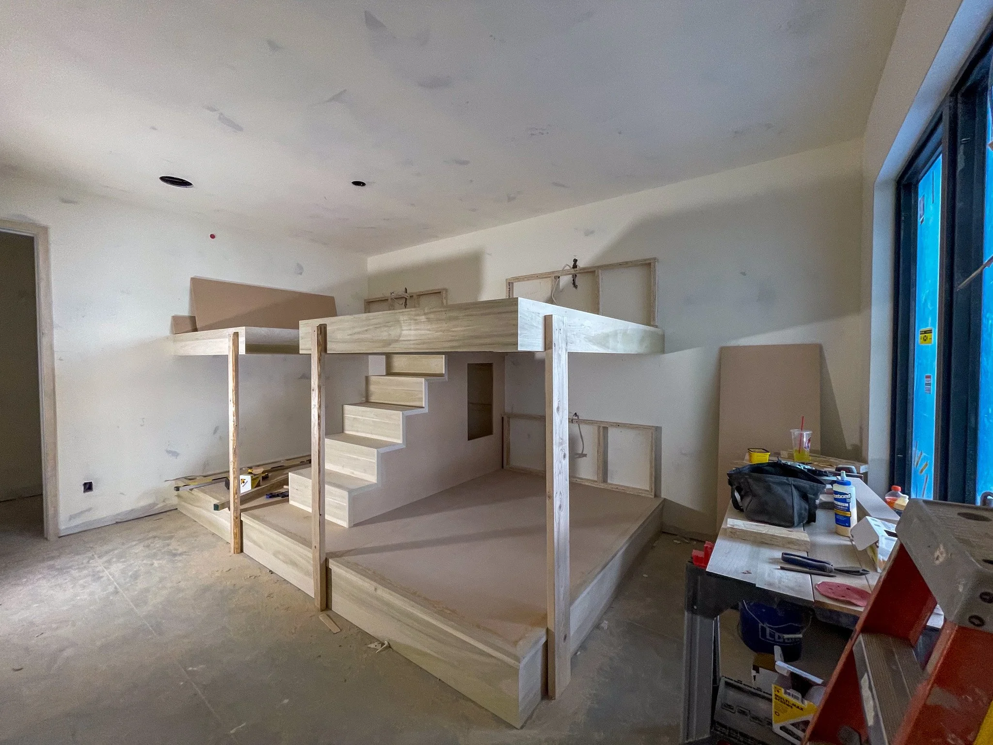 Interior of a room under construction with a built-in wooden bunk bed with stairs, open frame structures, and construction tools and materials on a work table.