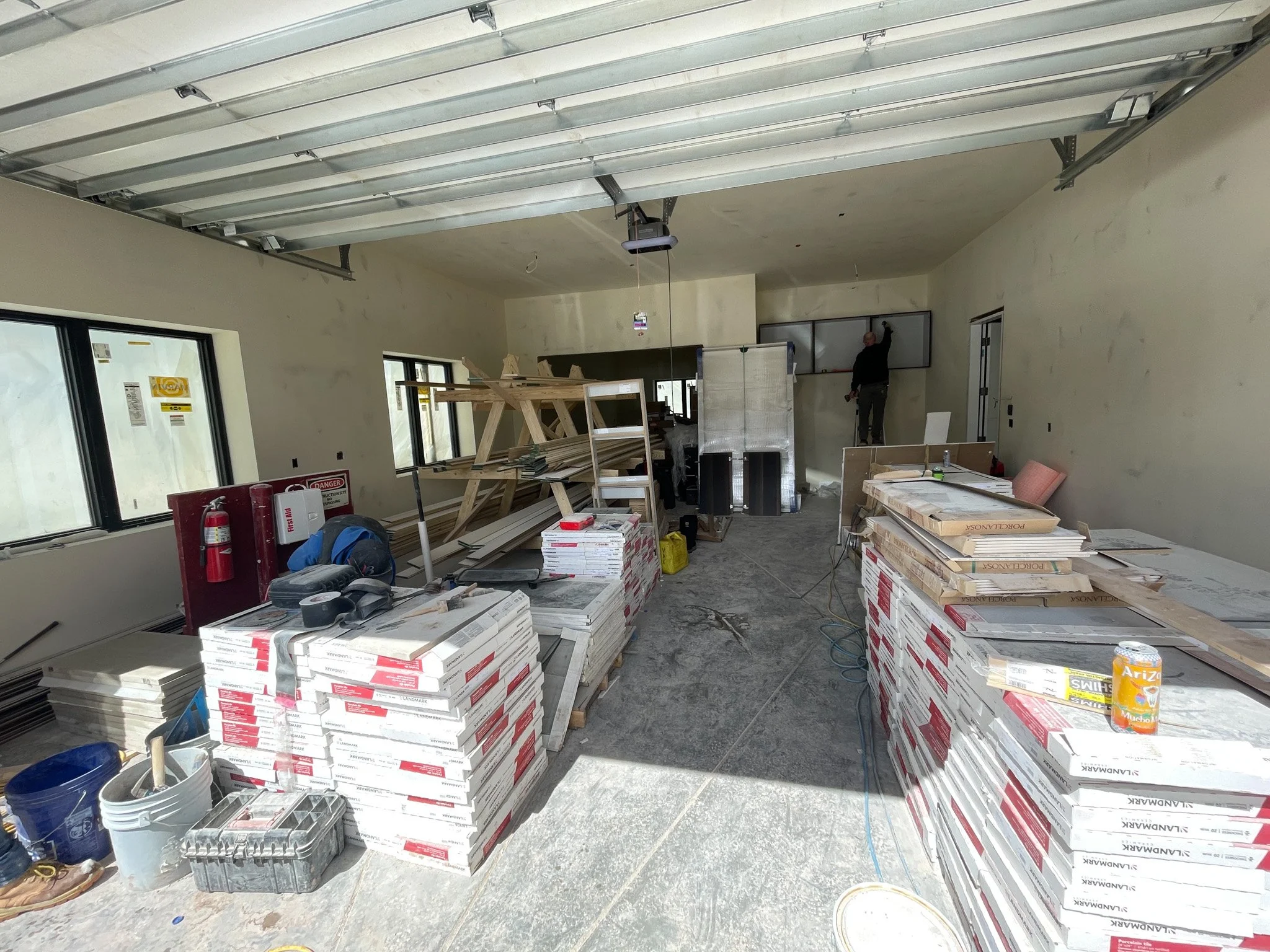 Interior of a house under construction with stacks of drywall, construction tools, and a worker on a ladder working on a wall.