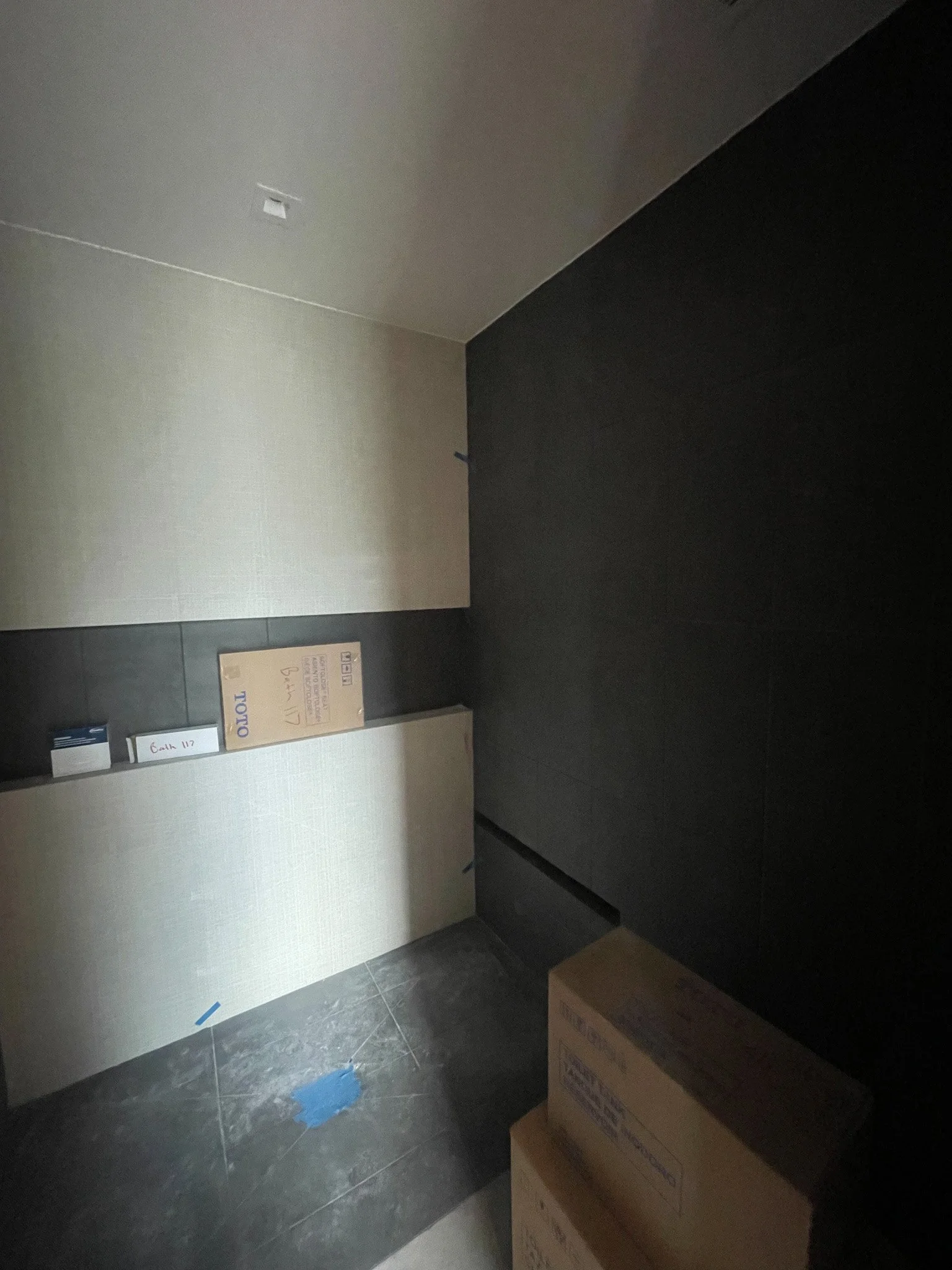 A room with dark flooring, beige wall panels, and a black wall. There are cartons and boxes stacked in the room, some sealed and others open.