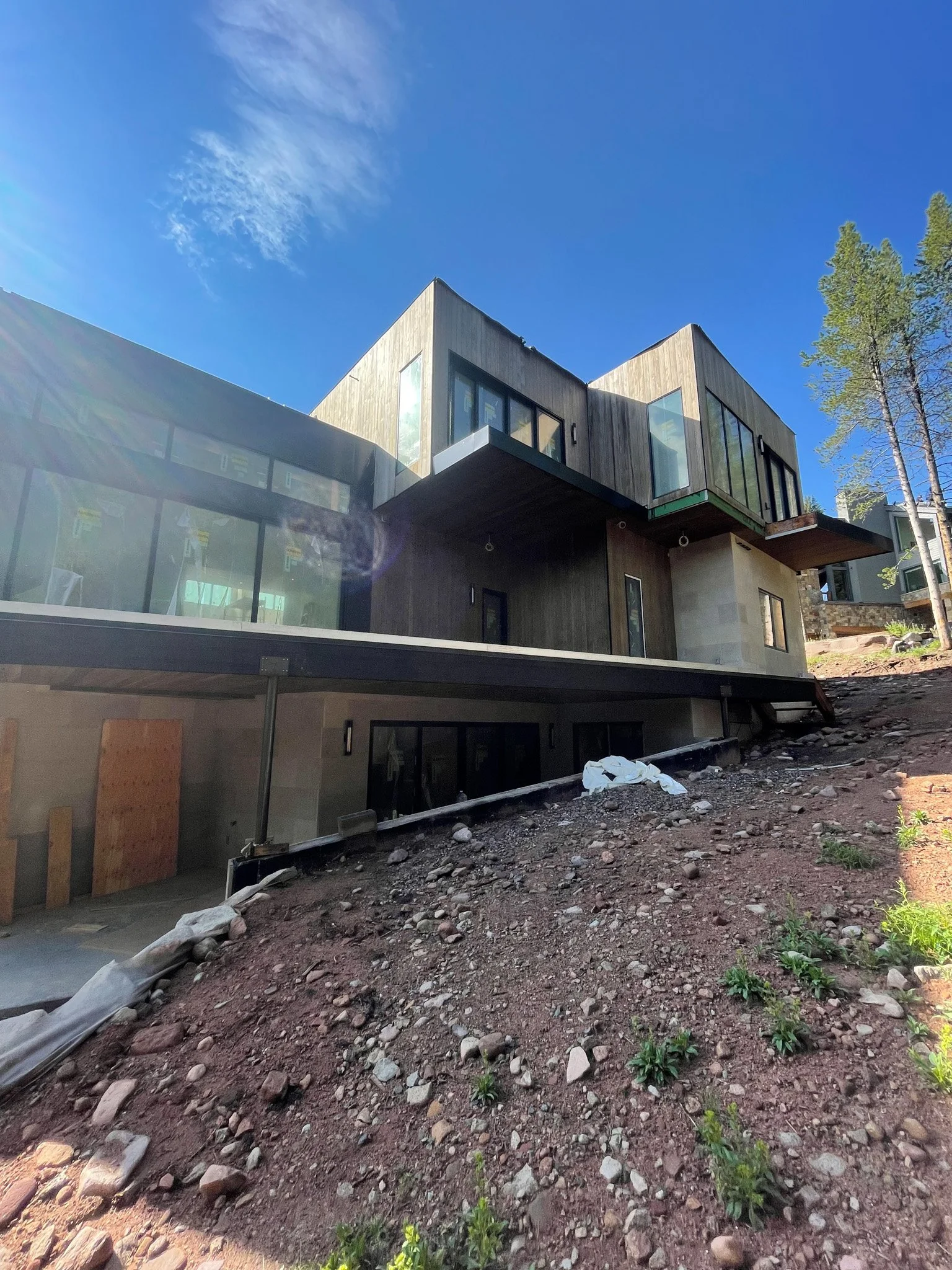 Modern multi-story house under construction on a hilly terrain, with wooden and glass exterior, surrounded by trees and a clear blue sky.