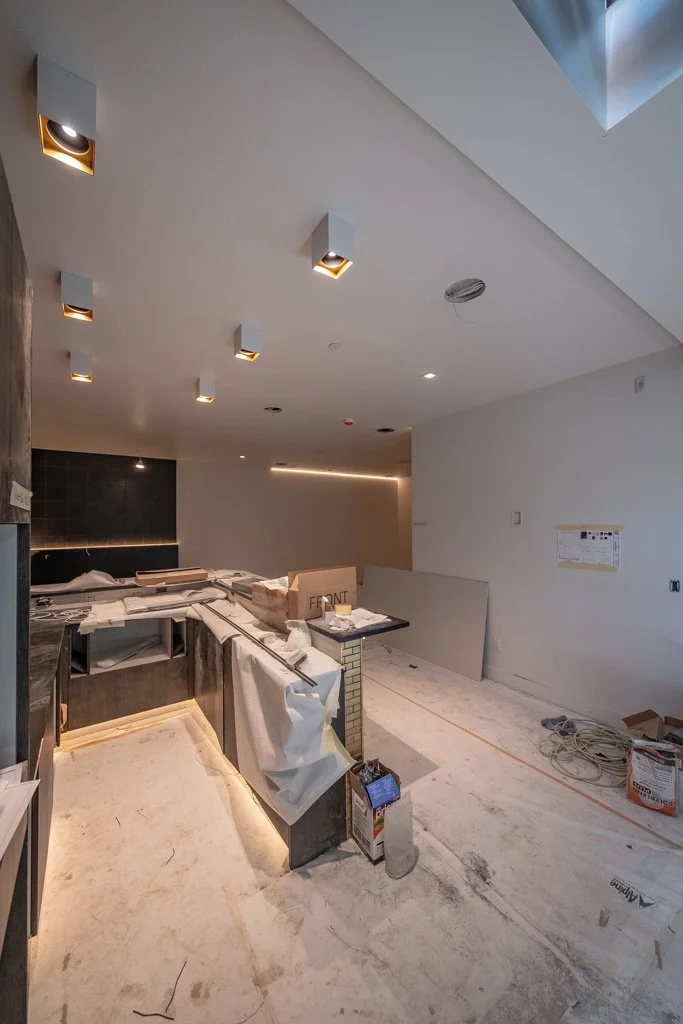 Under construction kitchen area with white walls, ceiling lights, and a black backsplash. Construction materials and tools are on the countertop and floor.
