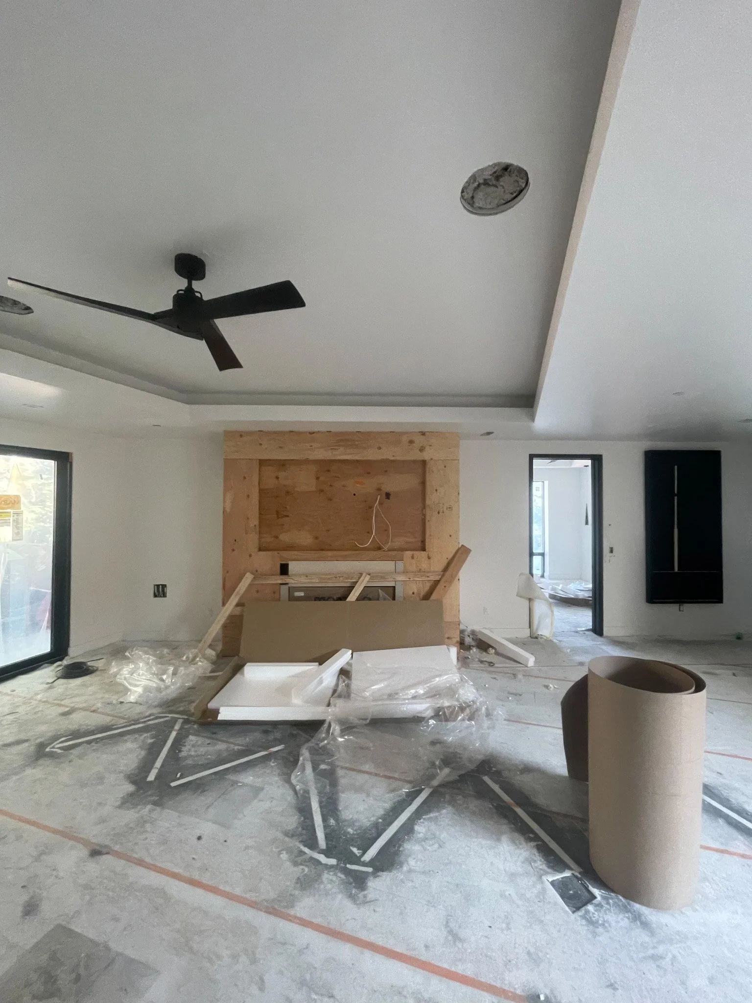 Interior of a room under construction with unfinished work, debris on the floor, a black ceiling fan, and a large window.