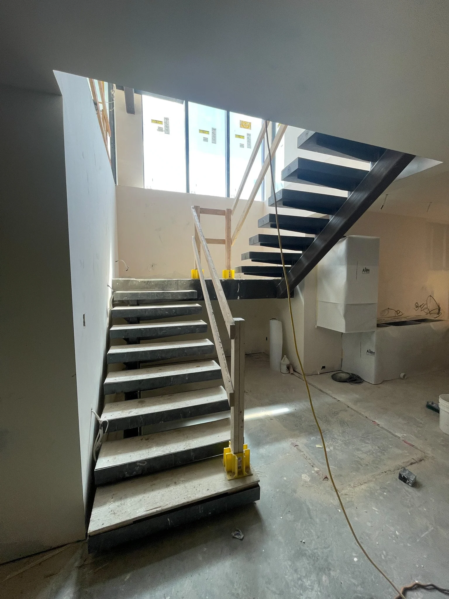 Construction site interior with an unfinished staircase, black floating steps, and temporary yellow supports, under natural light from large windows.
