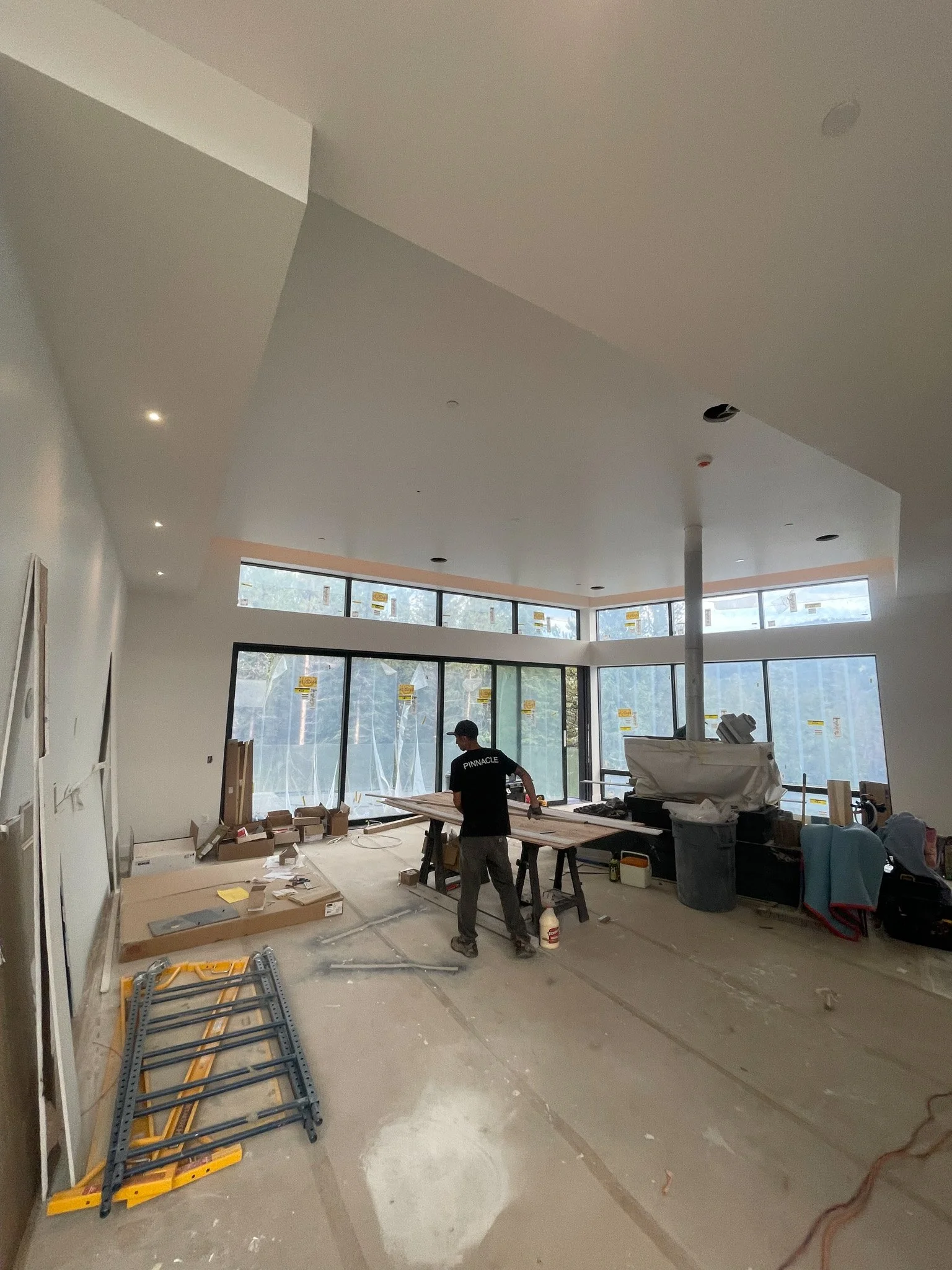 Interior space under construction with a worker cutting wood at a worktable, large windows at the far end, construction tools and materials scattered on the floor.