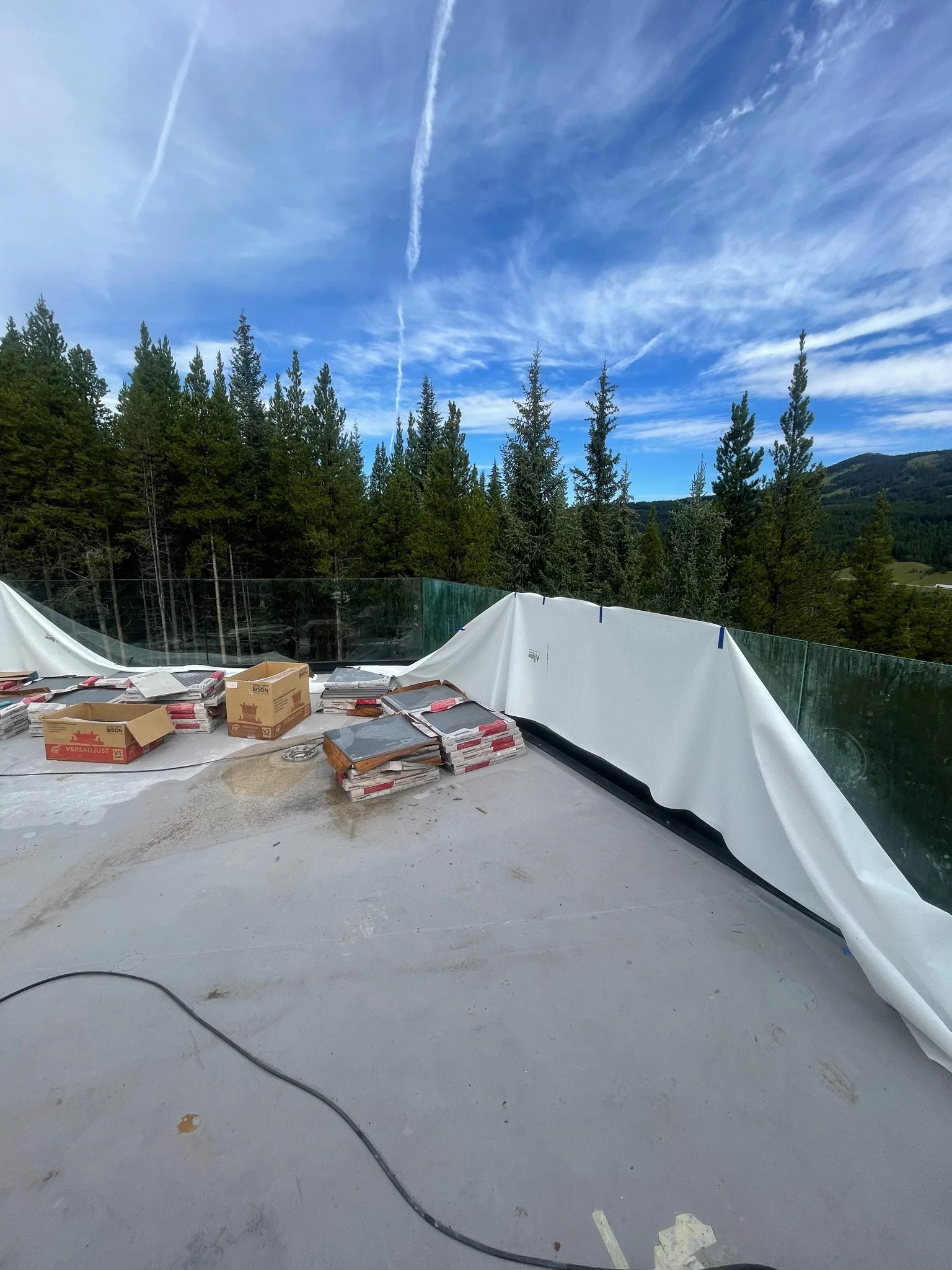 View of a construction site or unfinished balcony with stacks of tiles and supplies, surrounded by protective plastic and glass barriers, with a background of green trees and a partly cloudy blue sky.