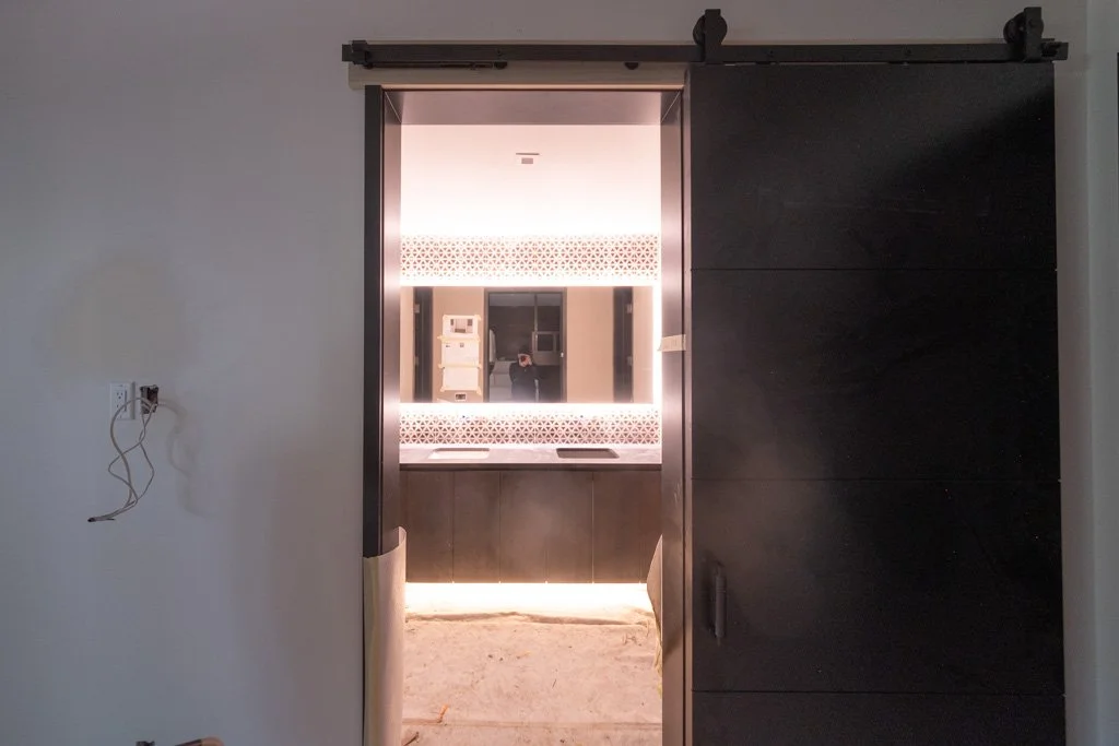 Black sliding barn door open to reveal a modern bathroom with a large mirror, patterned wall tiles, and a vanity with lighting underneath.