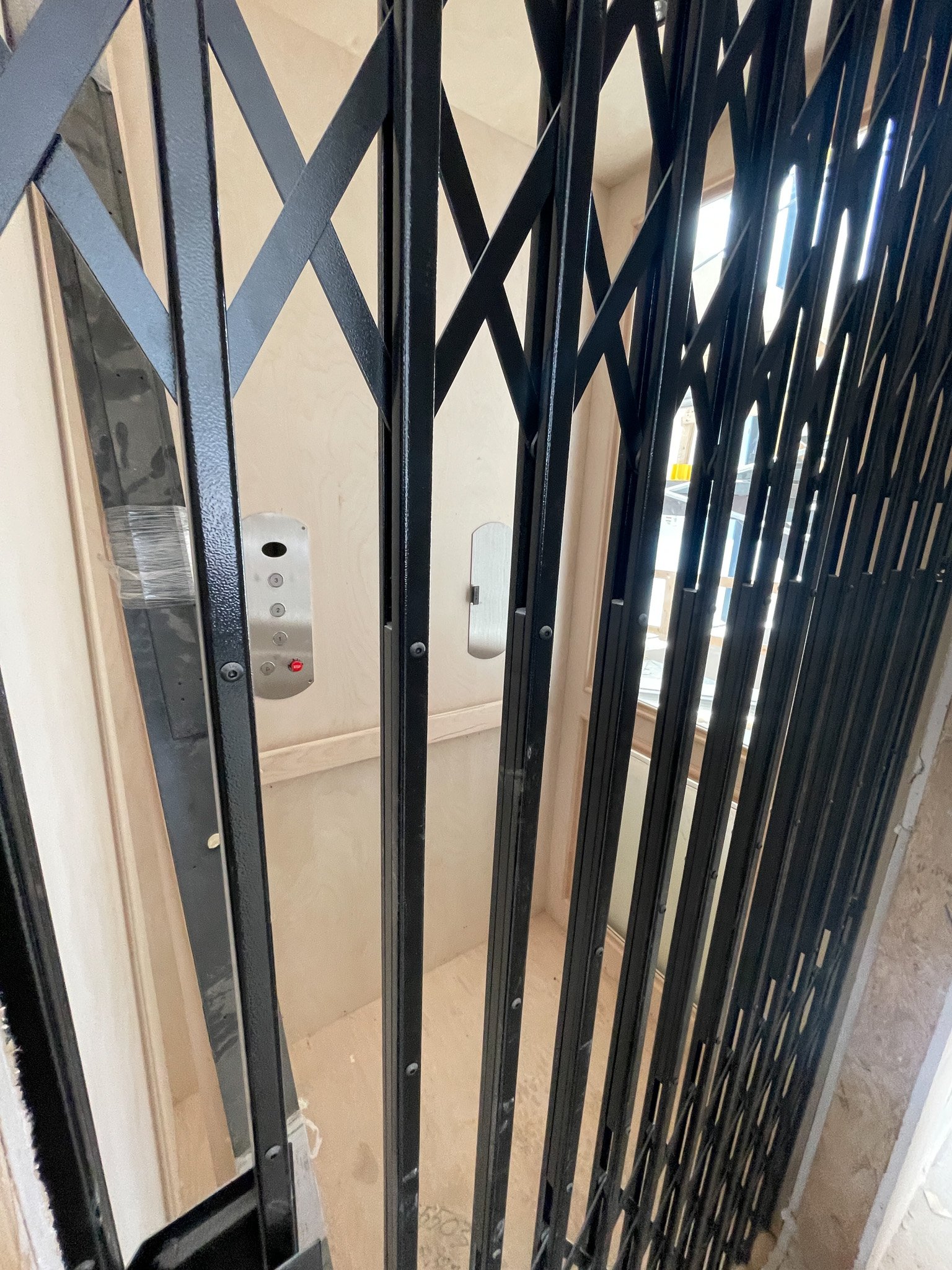 Inside view of an elevator with black safety gate, button panel on the wall, and a window allowing sunlight to enter.