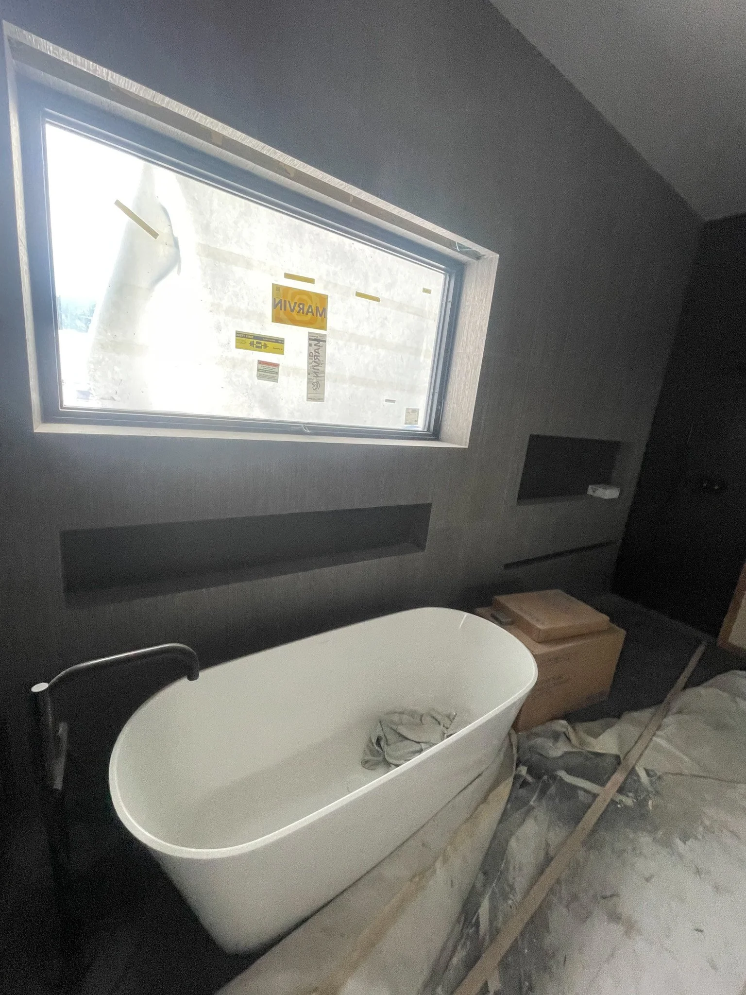 Bathroom under construction with a white bathtub, a window marked 'Marvin,' a cardboard box, and construction materials on the floor.